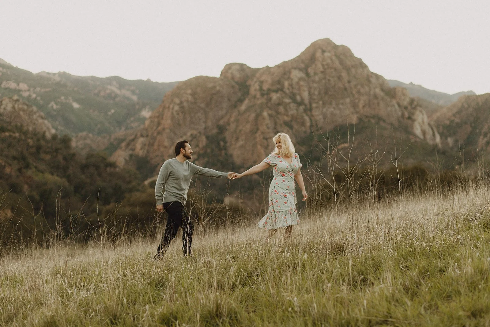 A man and a woman holding hands in a grassy field with mountains in the background.