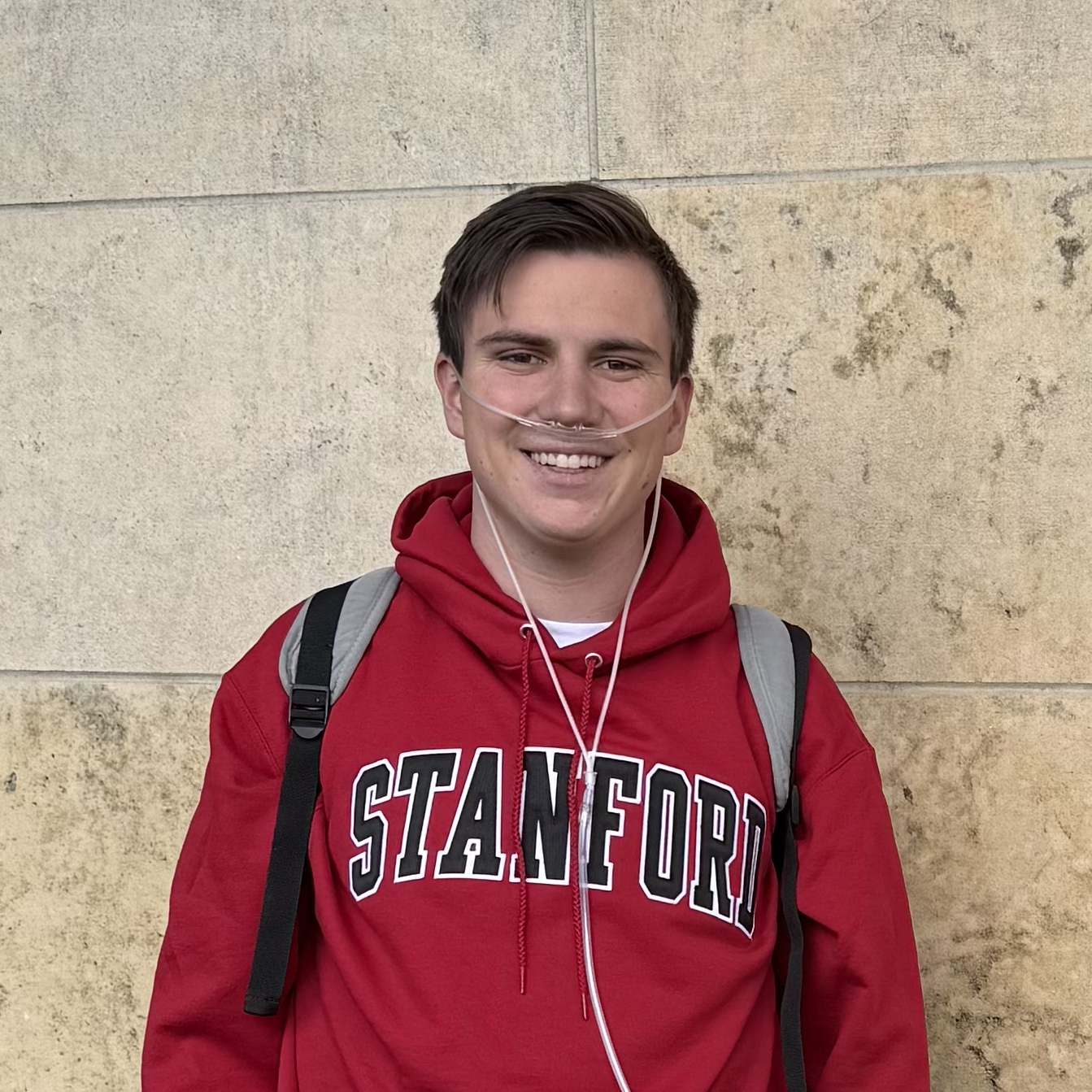 A young man smiling outdoors wearing a red Stanford sweatshirt, a gray backpack, and a nasal oxygen tube, standing against a beige stone wall.