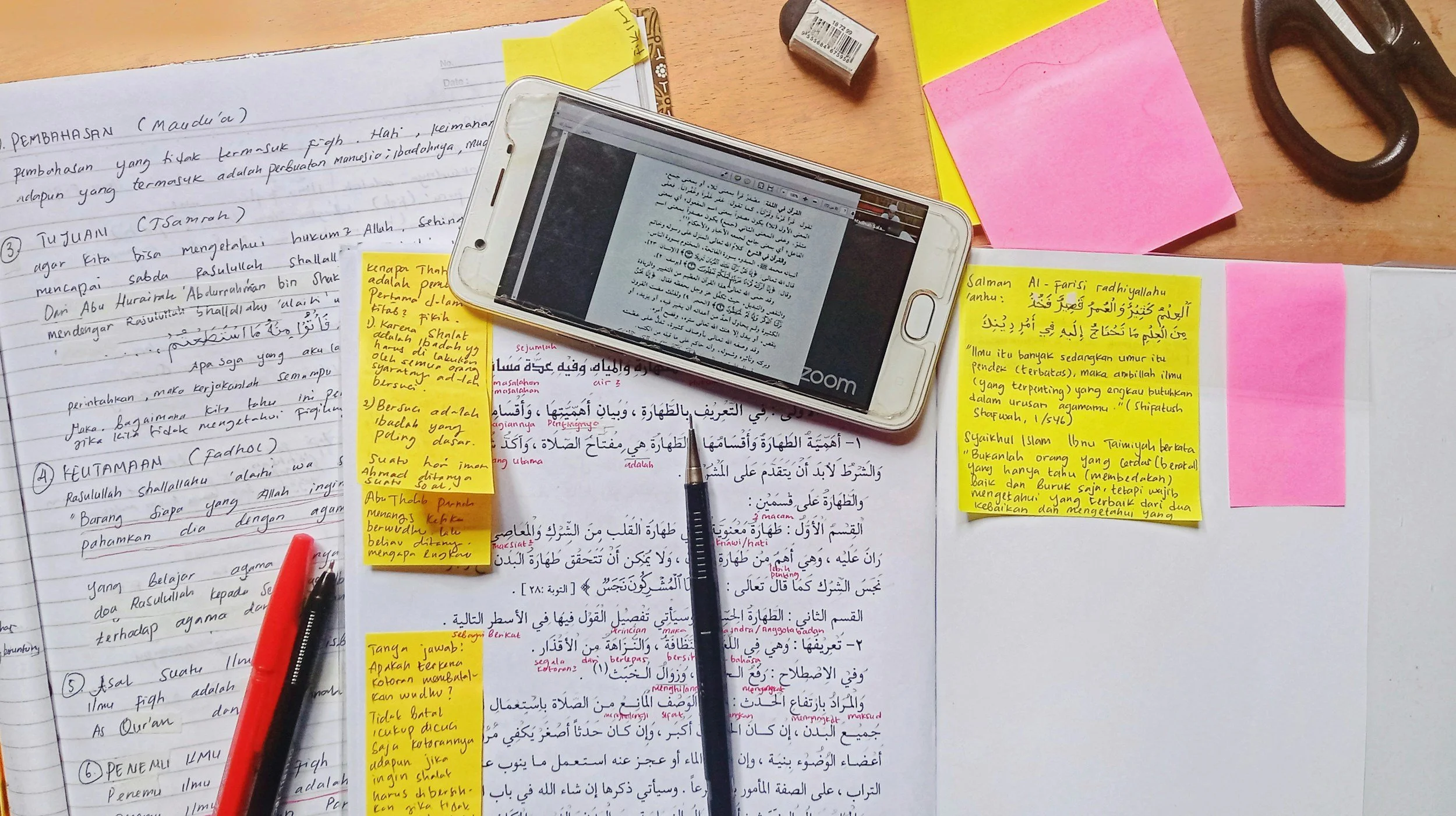 A study desk with handwritten notes, yellow sticky notes and pink sticky notes, a smartphone with a video call, a pen, a pair of scissors, an eraser, and a yellow notebook.