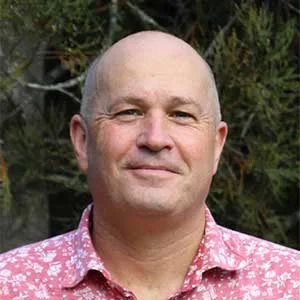 A middle-aged man with a bald head smiling outdoors with trees in the background, wearing a pink patterned shirt.