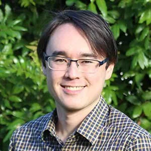 A young man with dark hair, glasses, and a checkered shirt smiling outdoors with greenery in the background.