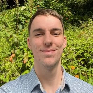 A young man with short brown hair smiling outdoors in front of green foliage and plants.