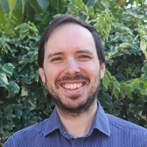 A smiling man with dark hair and a beard, wearing a blue patterned shirt, standing outdoors in front of green foliage.