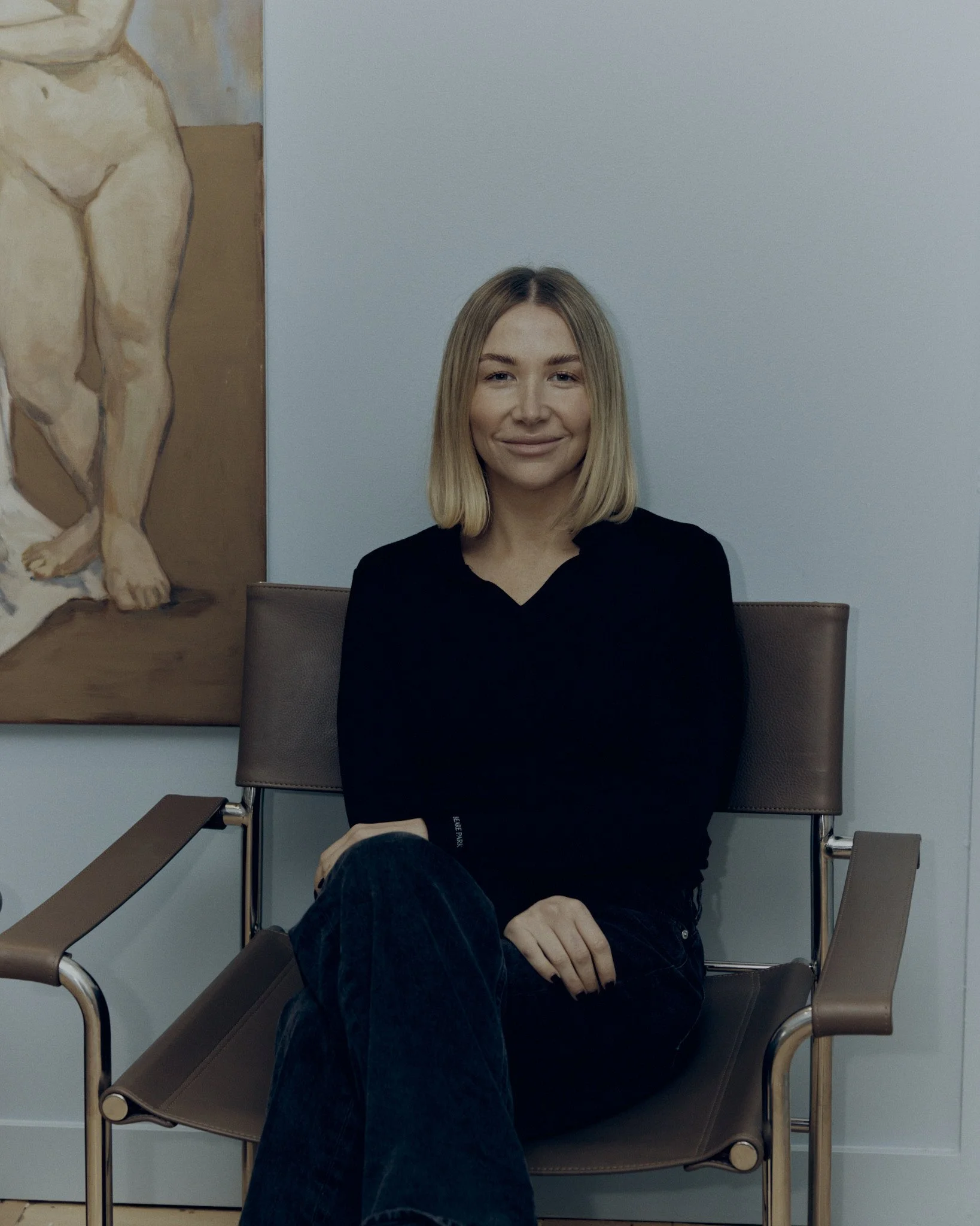 Young woman with blonde hair sitting on a brown chair in a room with light blue walls, with a painting of a lion in the background.