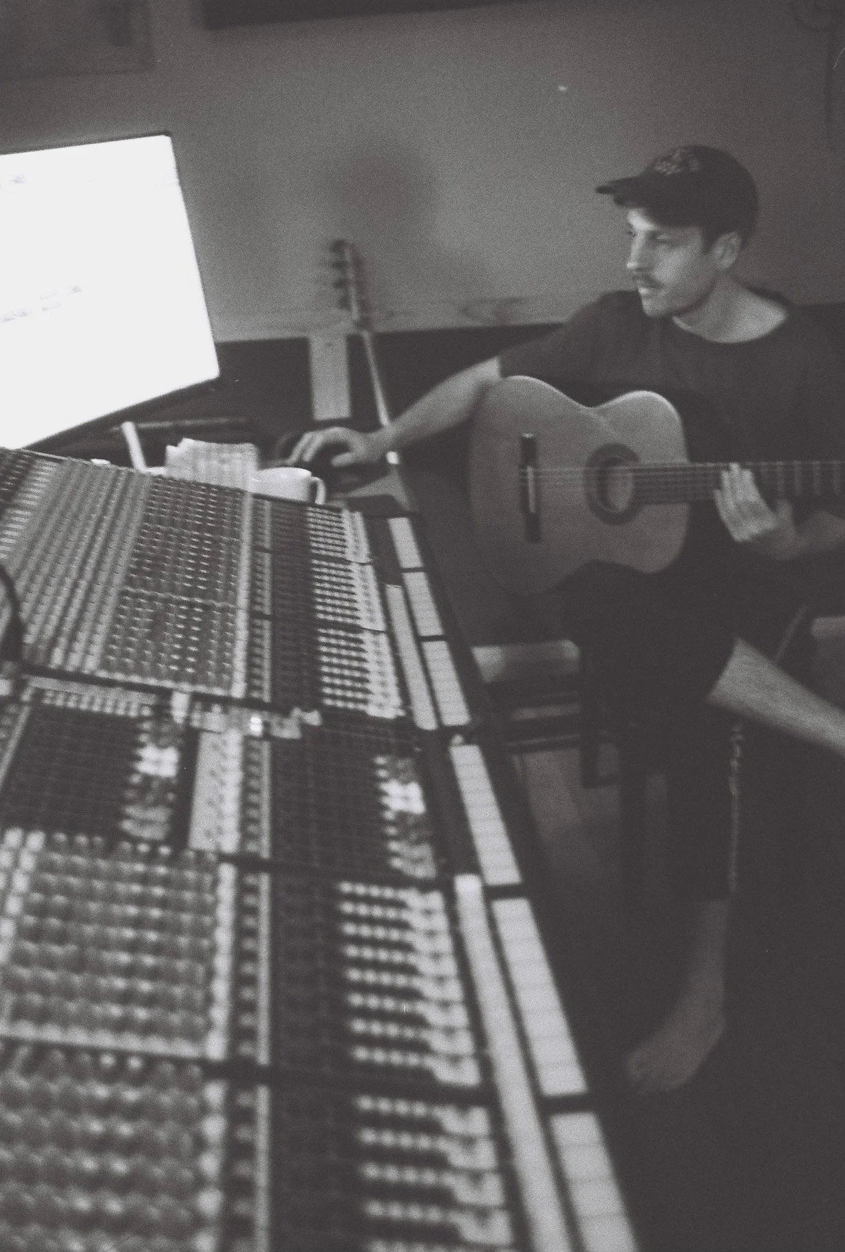 A person wearing a cap playing an acoustic guitar in a music studio, with a large mixing console and a computer monitor in front of them.