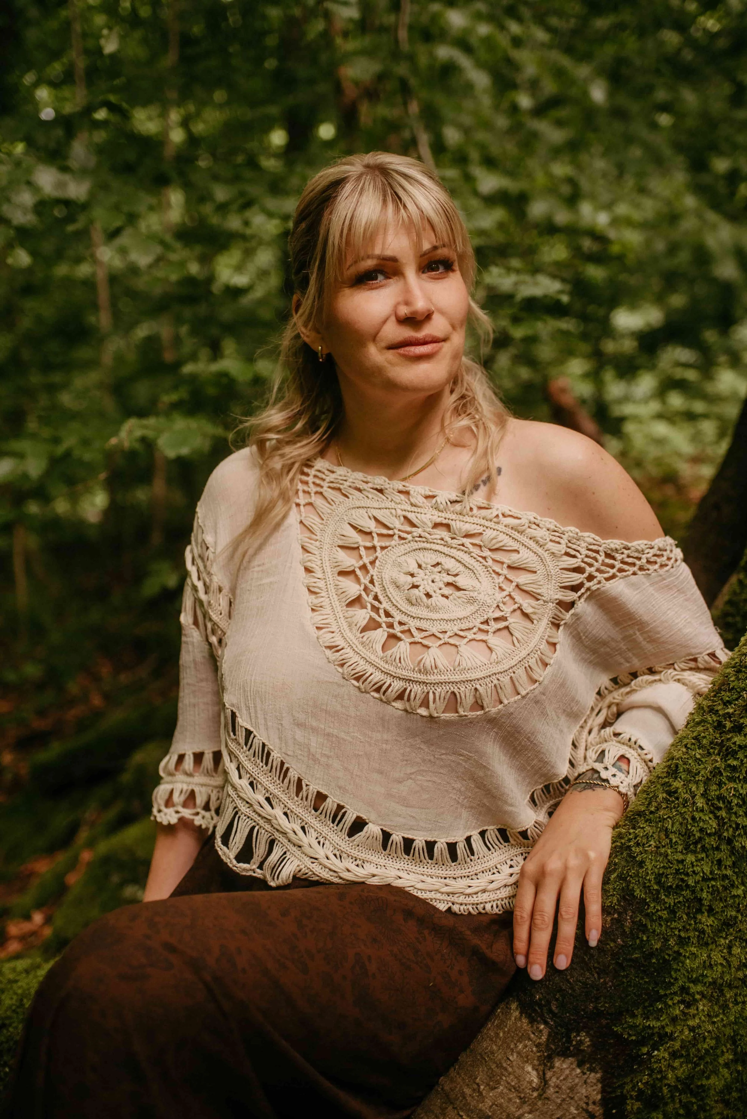 Anica von ANIMAcode sitzt draußen auf einem moosbedeckten Baumstamm im Wald, trägt beiges Häkeloberteil und braune Hose, symbolisiert Ruhe, emotionale Balance und Achtsamkeit.