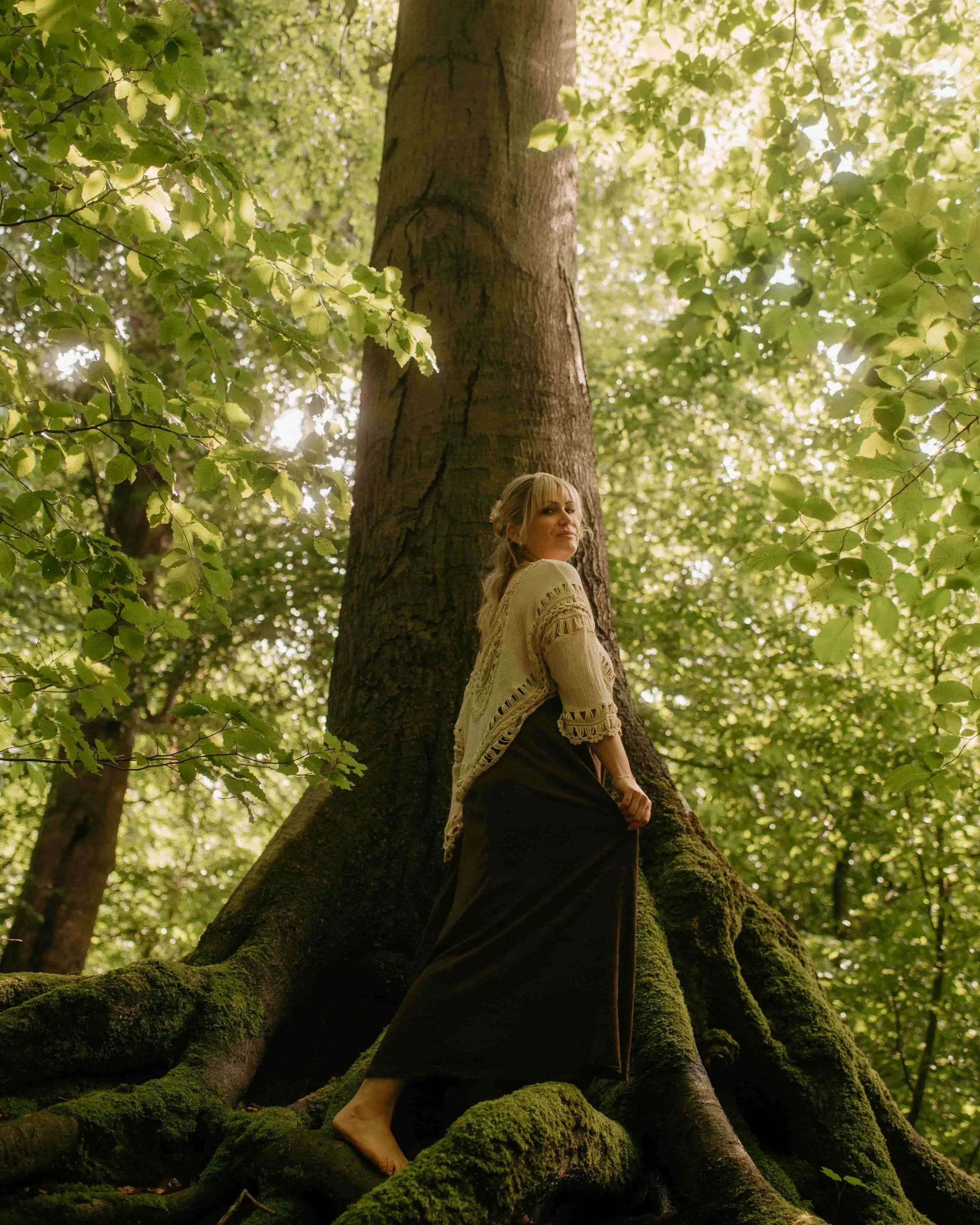 Frau in beigem Häkelpullover und dunklem Rock steht barfuß auf moosbedeckten Wurzeln eines großen Baumes im grünen Wald, symbolisiert Ruhe, emotionale Balance und Naturverbundenheit.