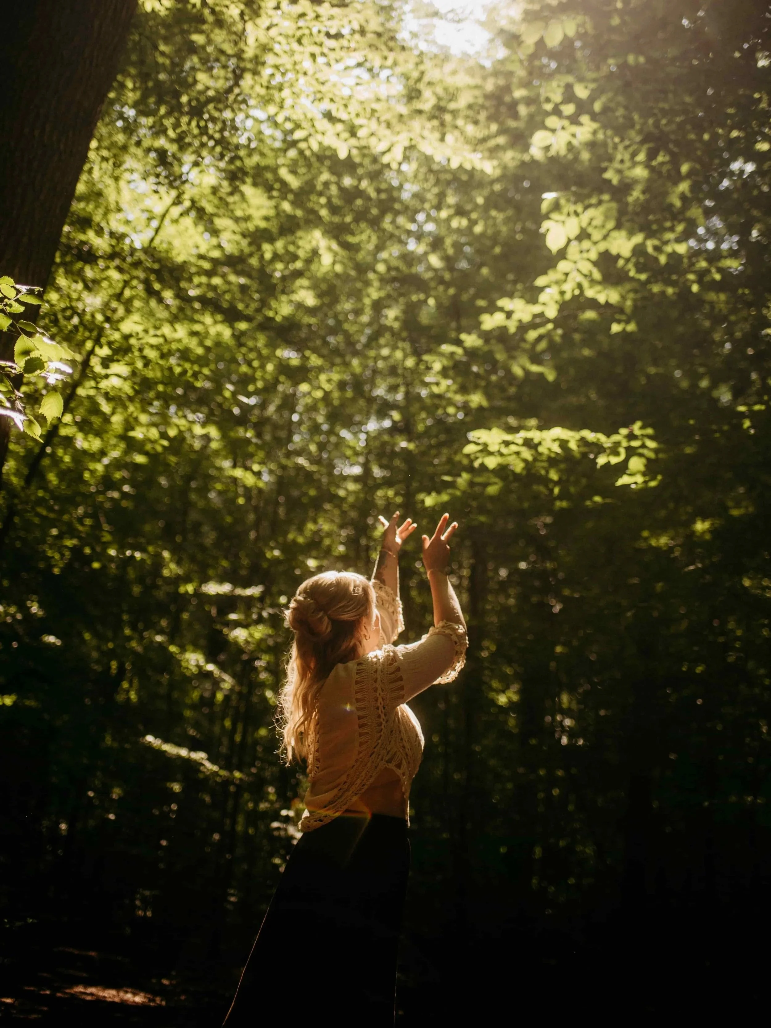 Blonde Frau steht in einem sonnendurchfluteten Wald, reflektierend und ruhig, Symbol für emotionale Balance und Wohlbefinden.