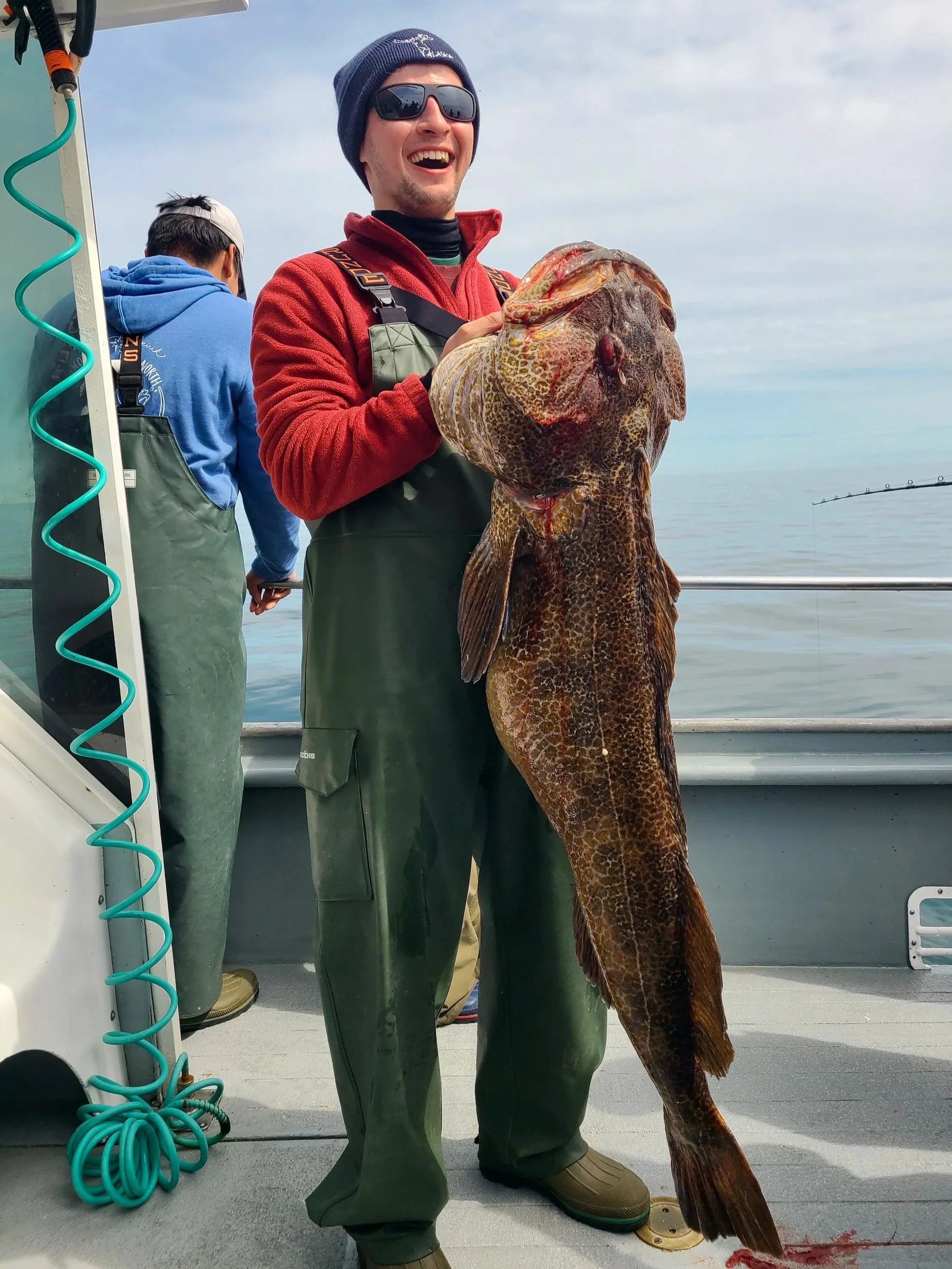 A man on a boat holding a large fish he caught, smiling and wearing sunglasses, a navy beanie, a red jacket, and green waterproof overalls.