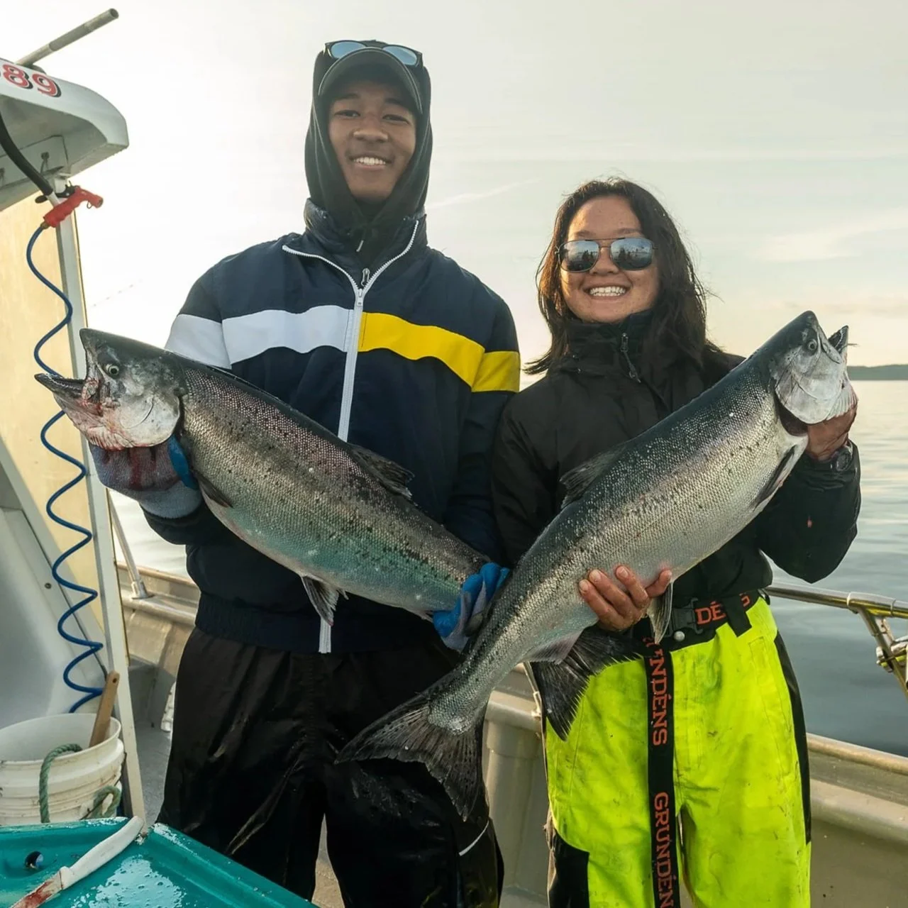 Two people on a boat holding large fish they caught, smiling at the camera, with water and a cloudy sky in the background.