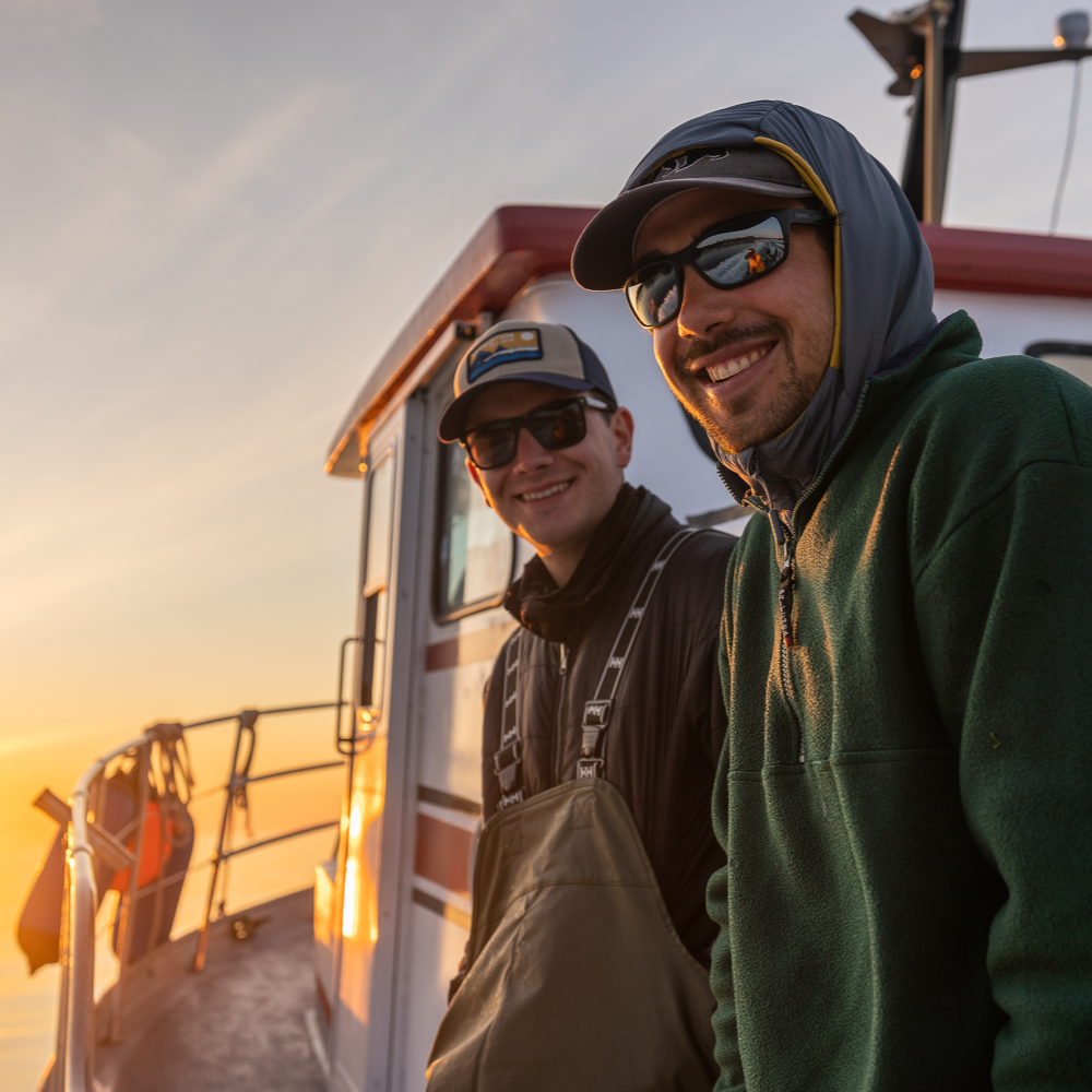 Two men on a boat during sunset, smiling and wearing sunglasses.