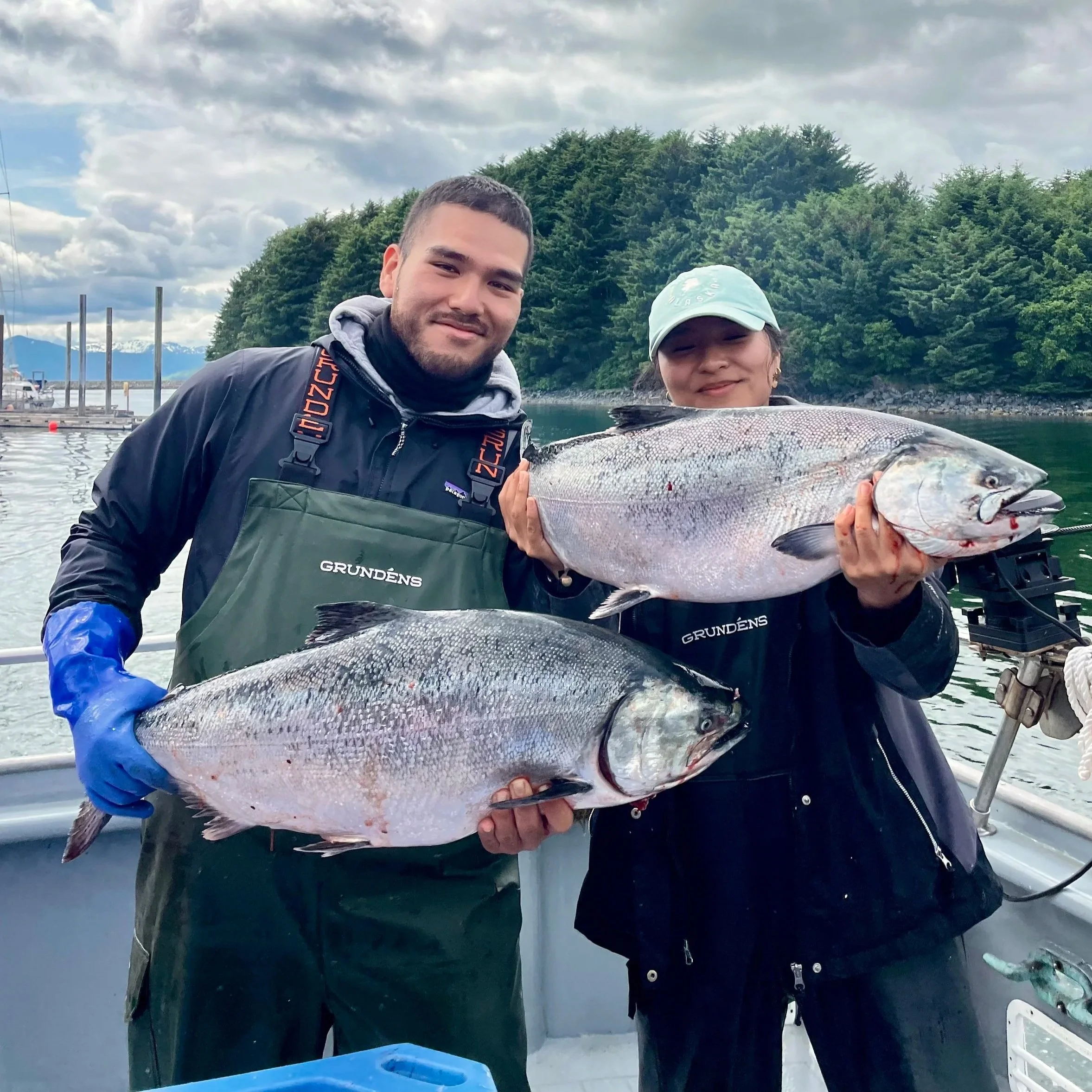 Two people on a boat holding large fish, with a backdrop of water, trees, and mountains under overcast skies.
