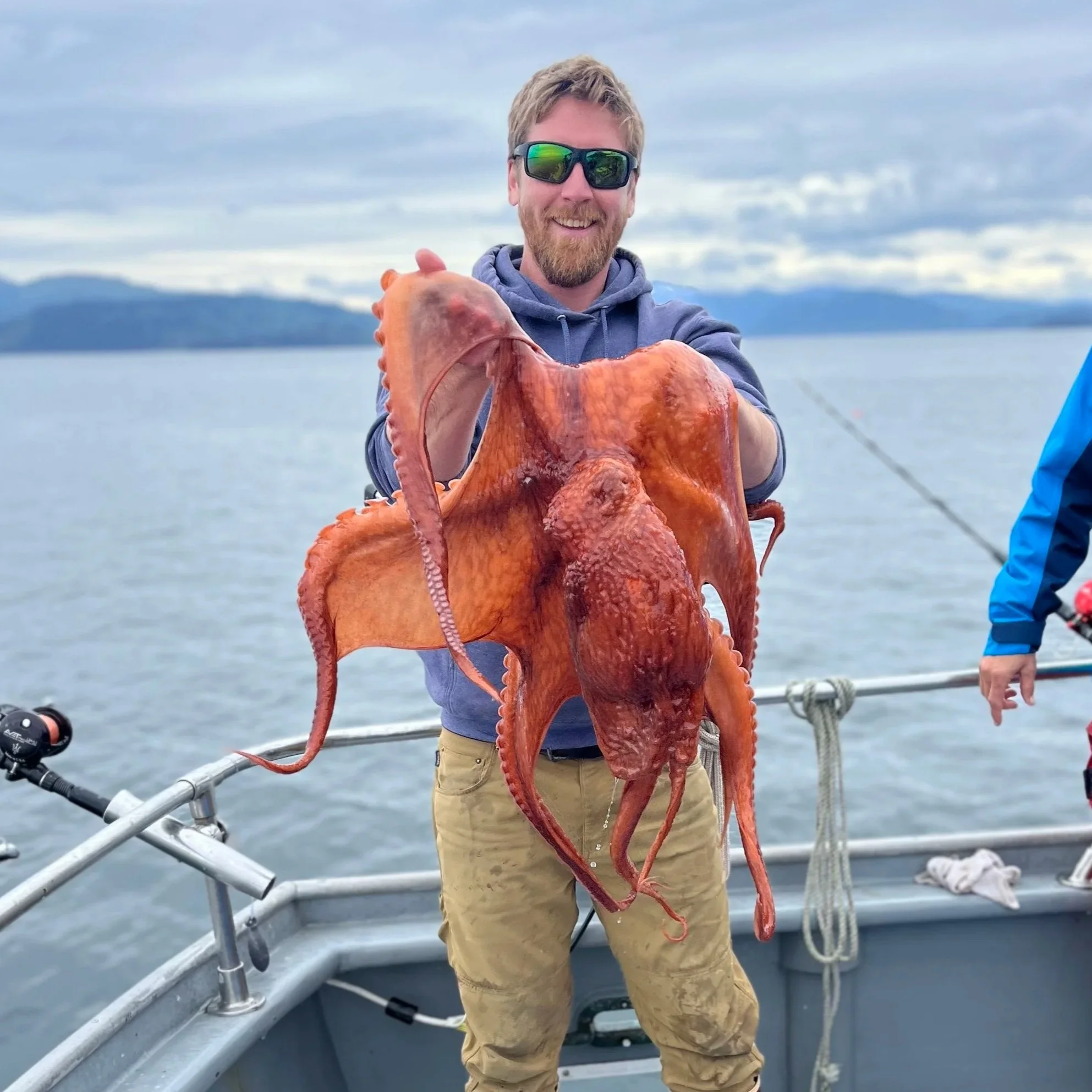 A man with sunglasses on a boat holding a large octopus, with water and distant land in the background.