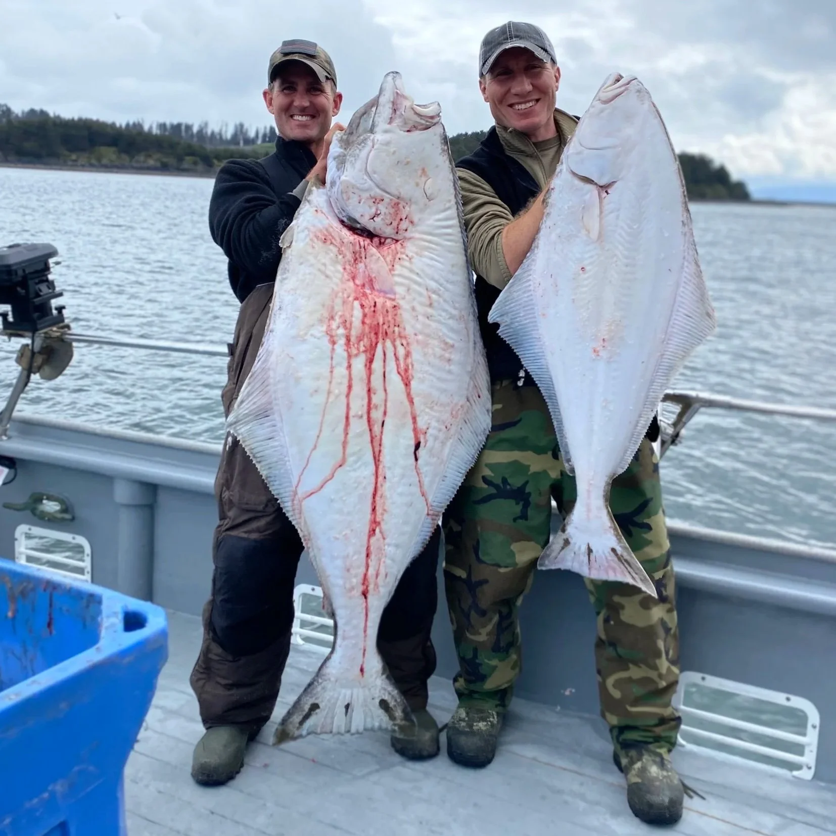 Two men holding large halibut fish on a boat in a body of water with cloudy sky.