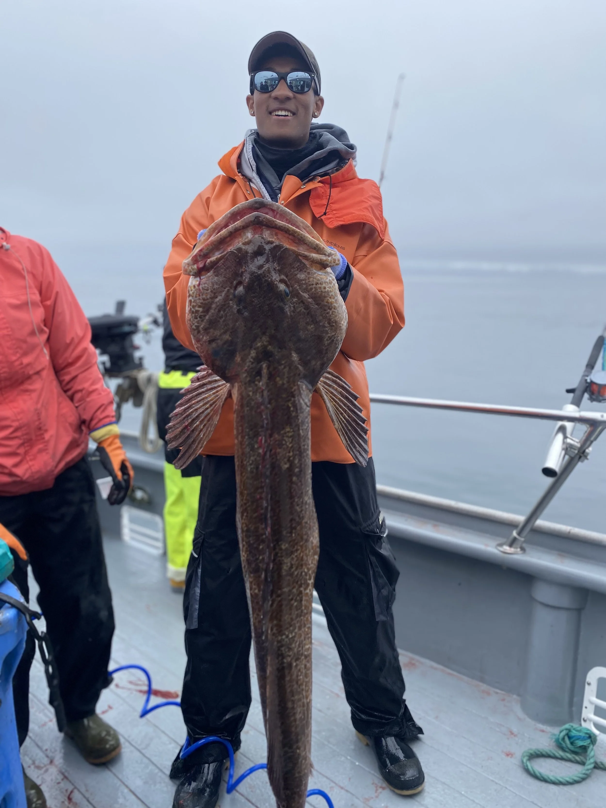 Person wearing an orange jacket, sunglasses, and a cap holding a large ling cod fish on a boat with others in the background.