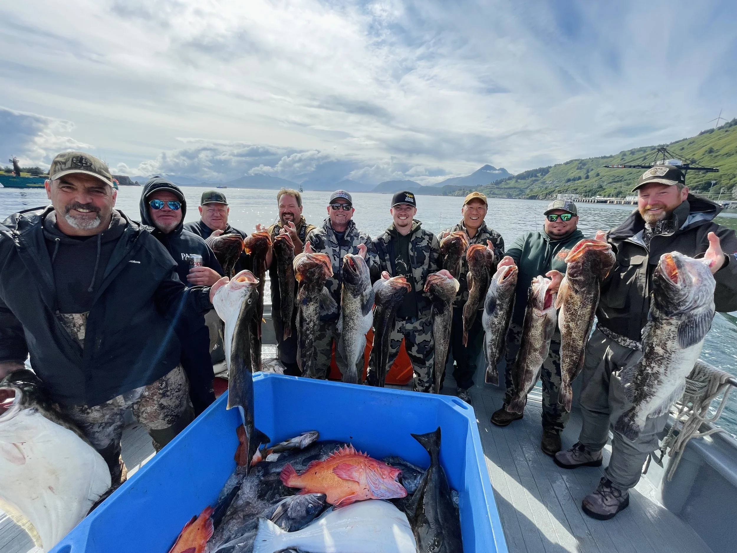A group of men on a boat holding up large fish, with a blue container filled with more fish in the foreground, and a scenic lake with mountains and cloudy sky in the background.