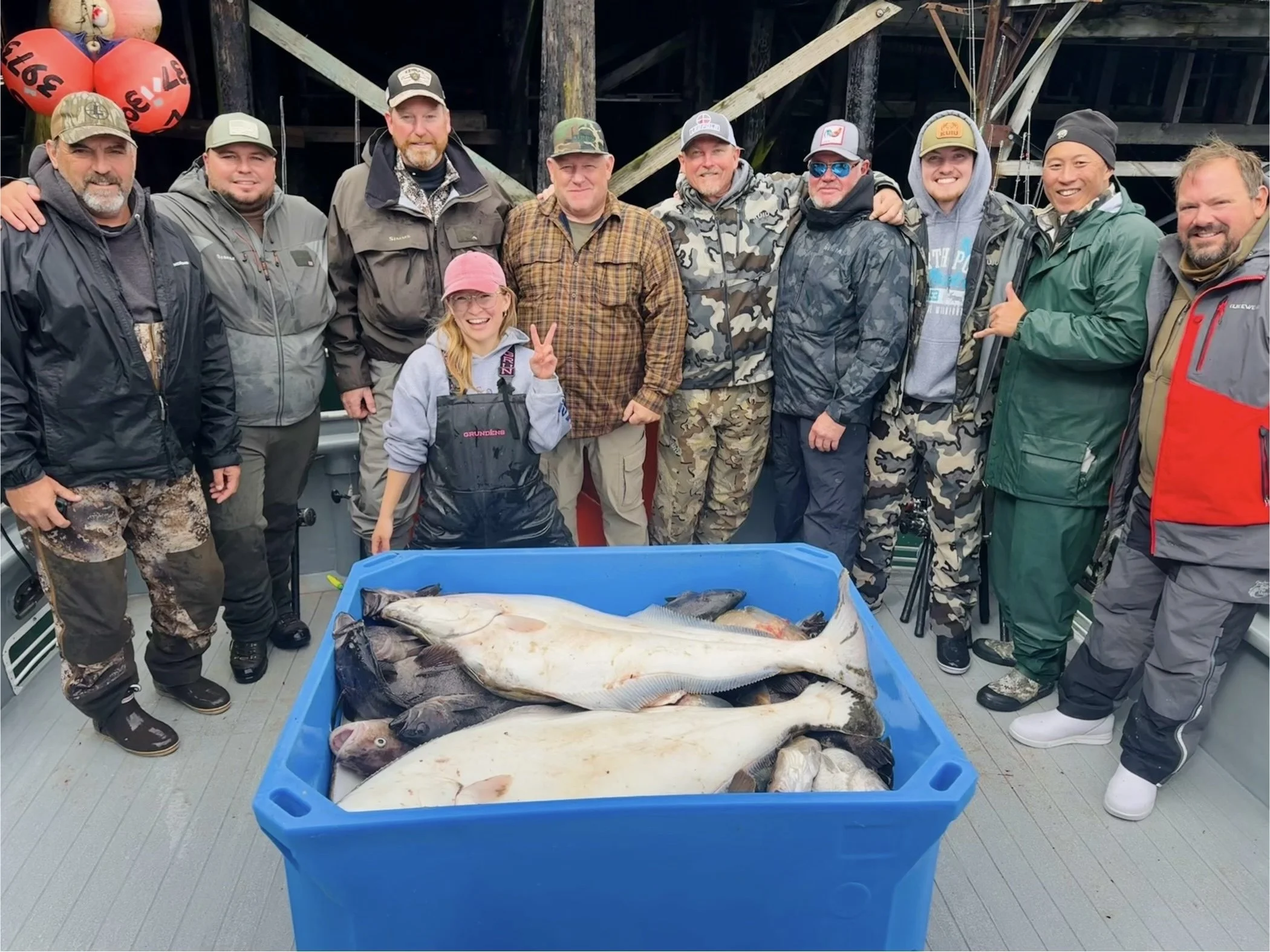 A group of people on a boat displaying a large catch of fish - king salmon, rockfish, ling cod, cod, halibut- with a girl in a pink cap flashing a peace sign and everyone smiling.