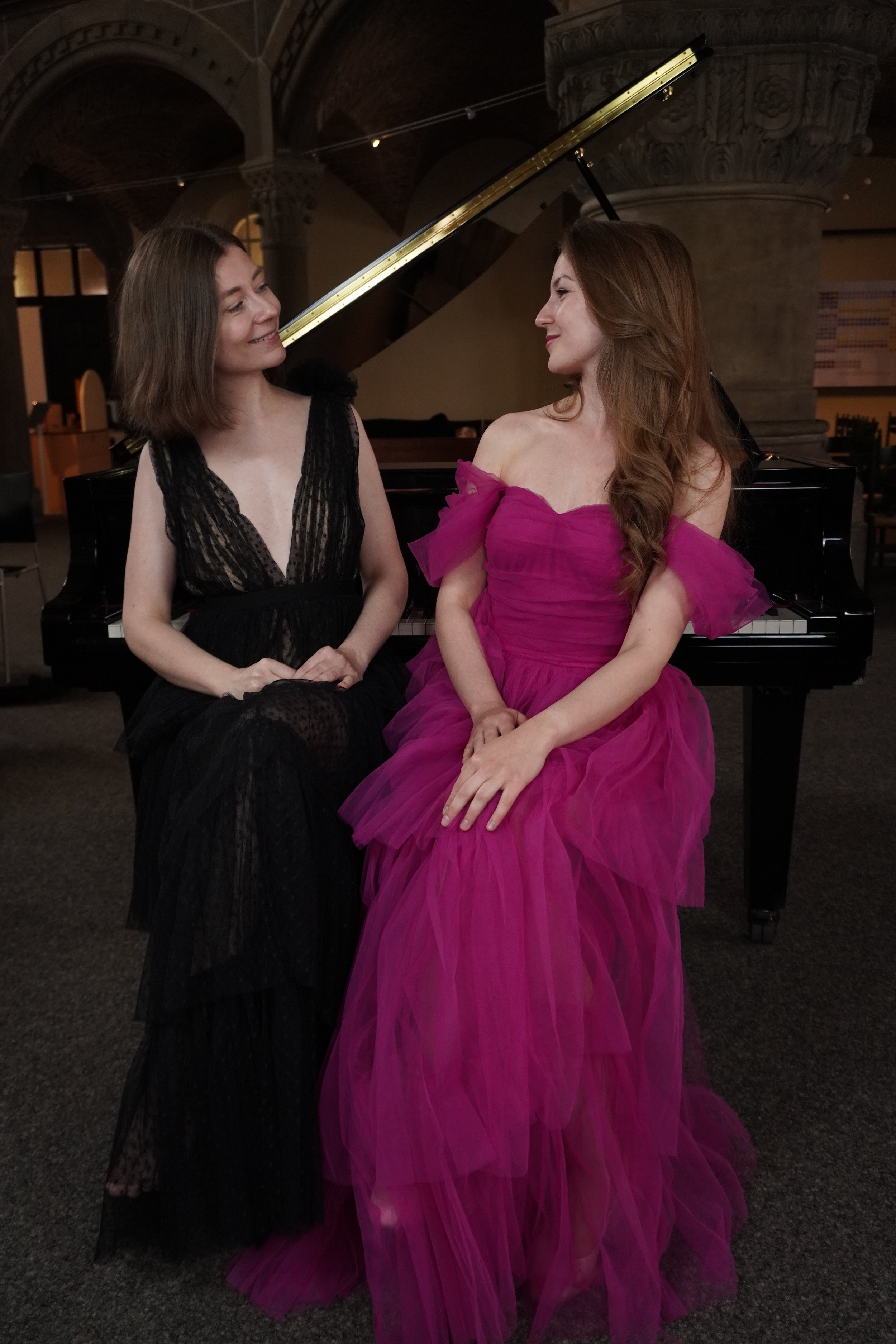 Zwei Frauen sitzen nebeneinander vor einem Klavier. Die Frau auf der linken trägt ein schwarzes, transparentes Kleid, die Frau auf der rechten trägt ein fuchsiafarbenes, voluminöses Tüllkleid. Beide schauen sich an und lächeln leicht.