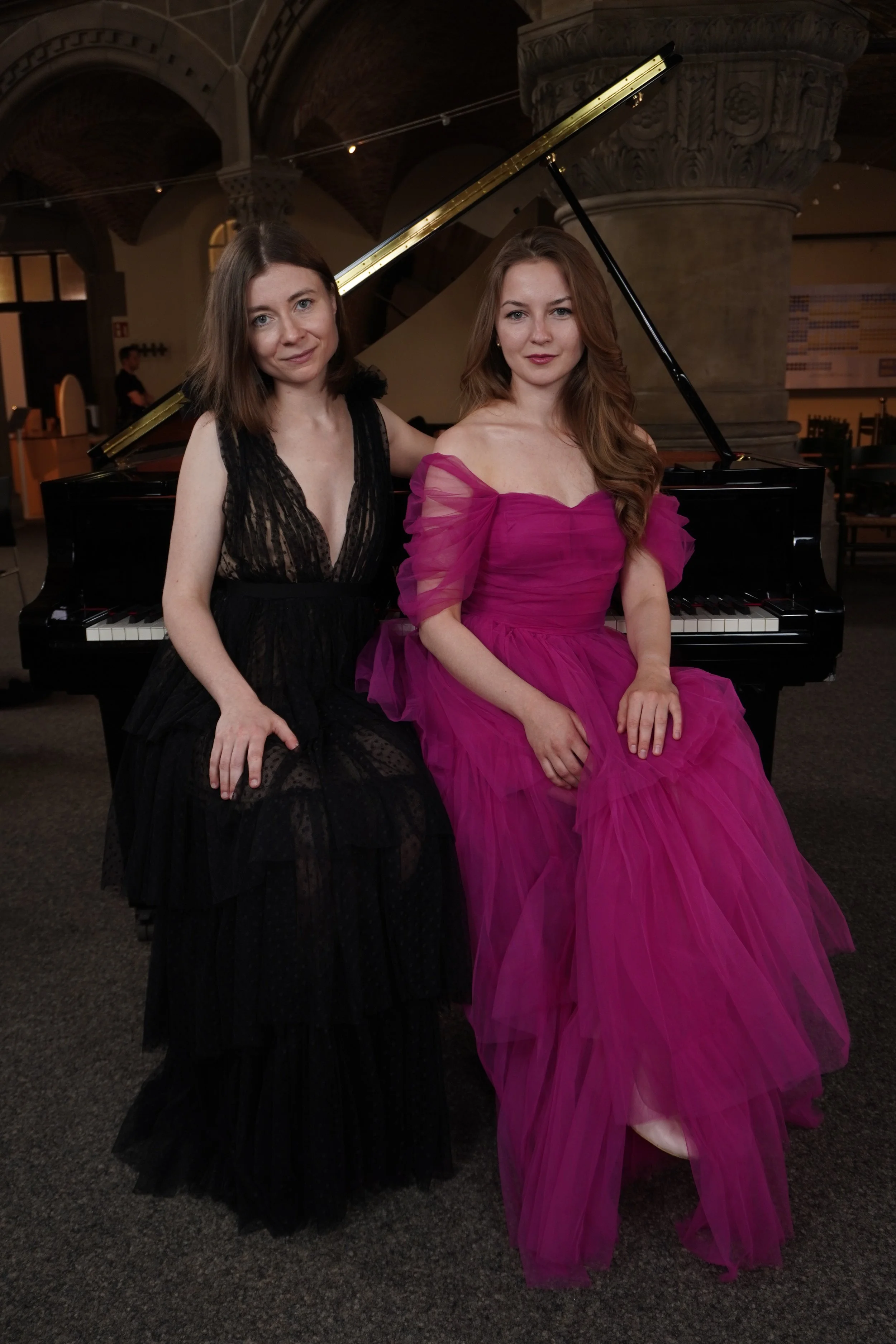 Zwei Frauen in eleganten Abendkleidern sitzen vor einem schwarzen Flügel in einem historischen Raum mit Steinarchitektur.