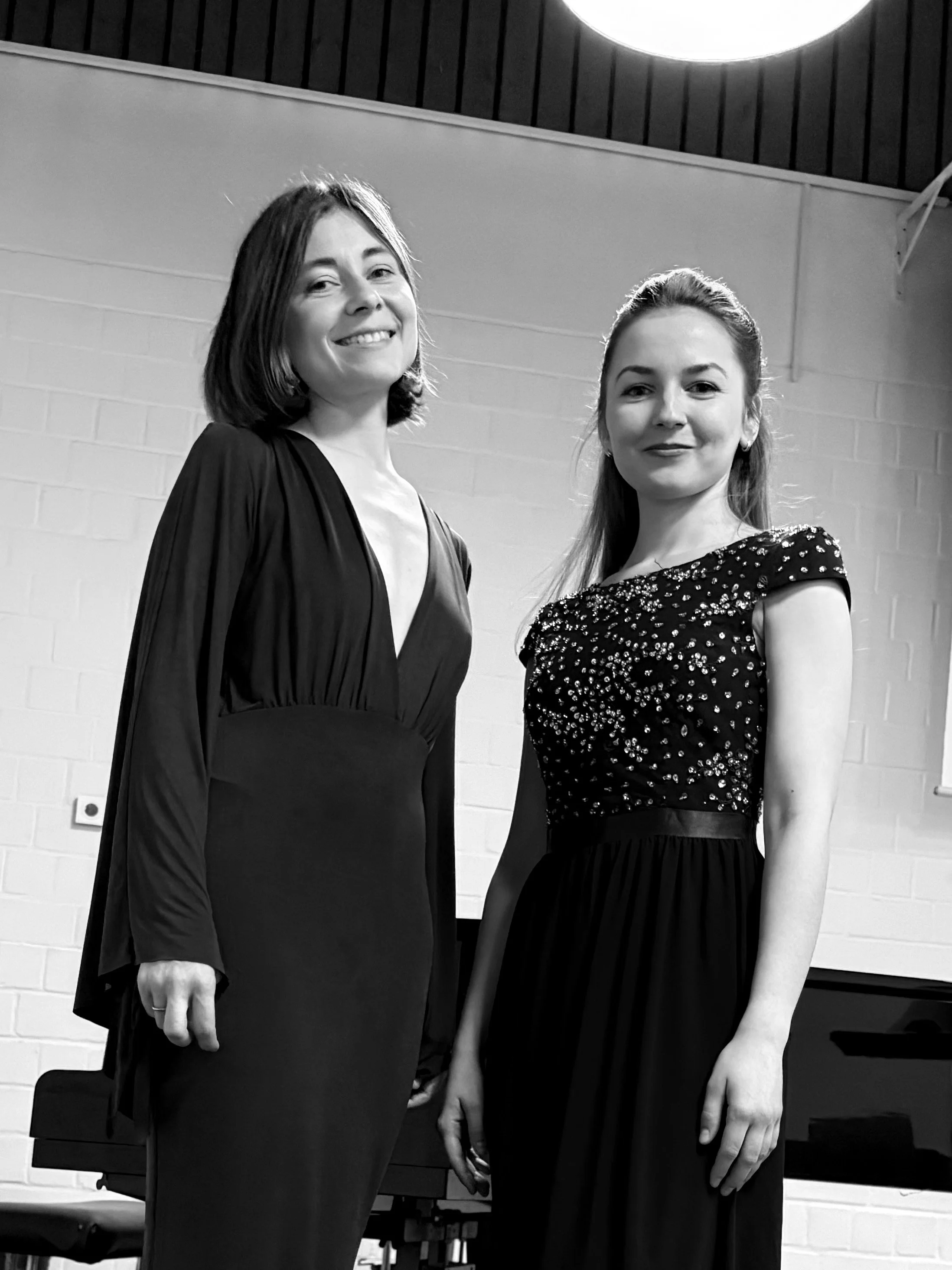 Zwei junge Frauen in eleganten Abendkleidern, die nebeneinander vor einer weißen Wand im Raum stehen.