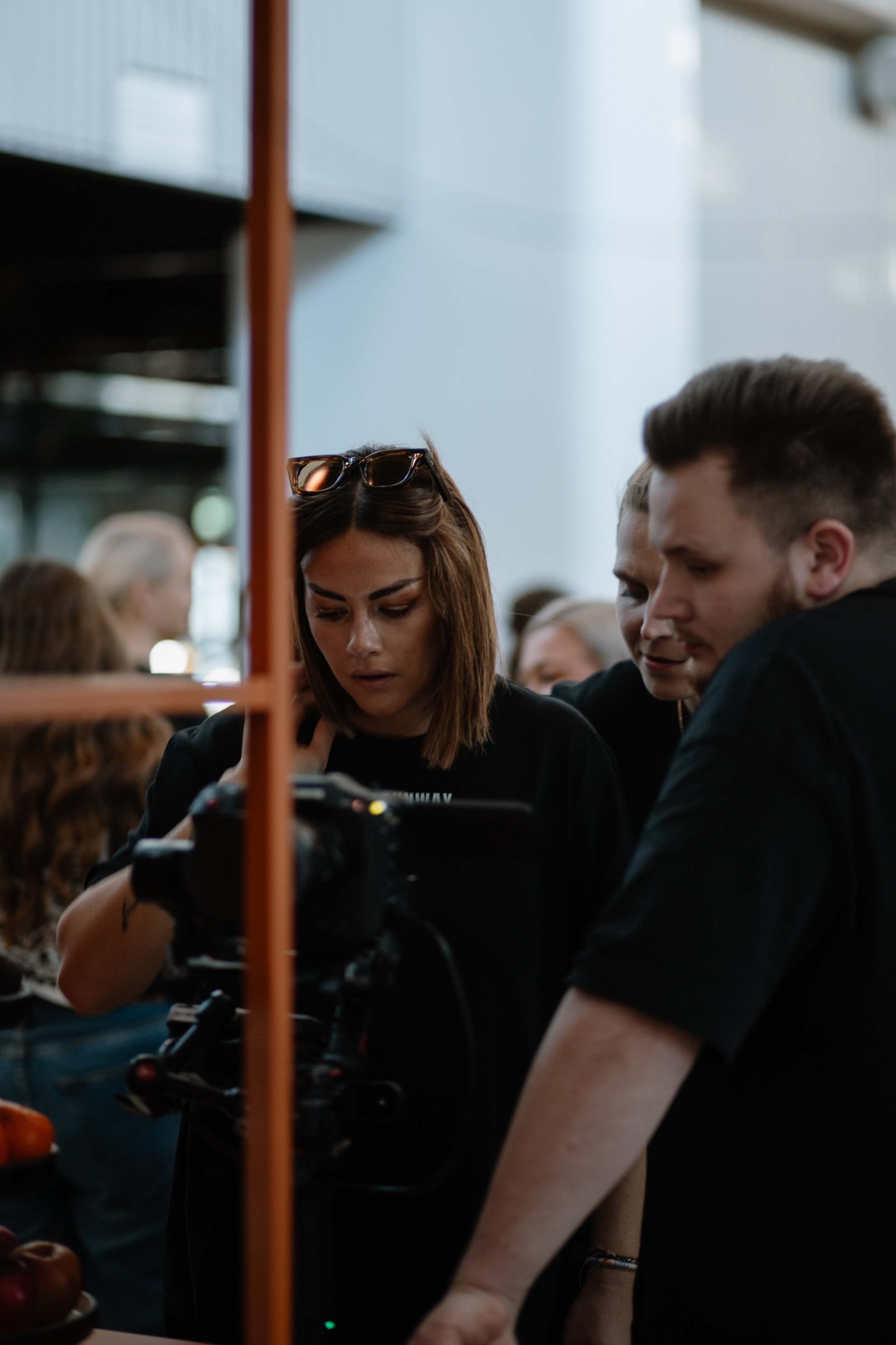 Three people look at a camera and a screen at an indoor event, with a woman in the center having sunglasses on her head and brown hair, a man to her right in a black shirt, and another man behind them also paying attention.