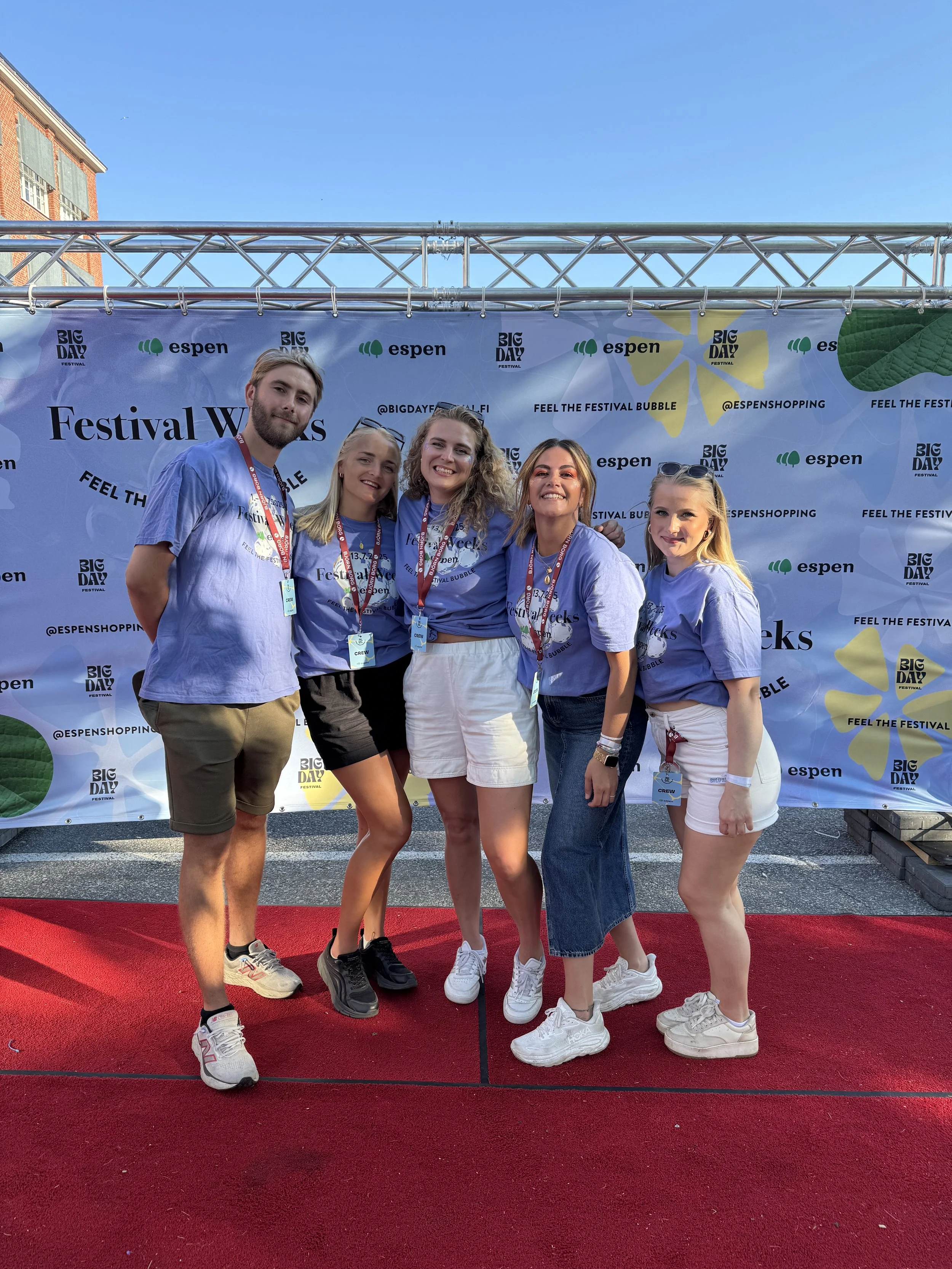 Group of five young people standing on a red carpet posing for a photo at a festival, all wearing matching blue T-shirts with festival branding and colorful lanyards, smiling with arms around each other.