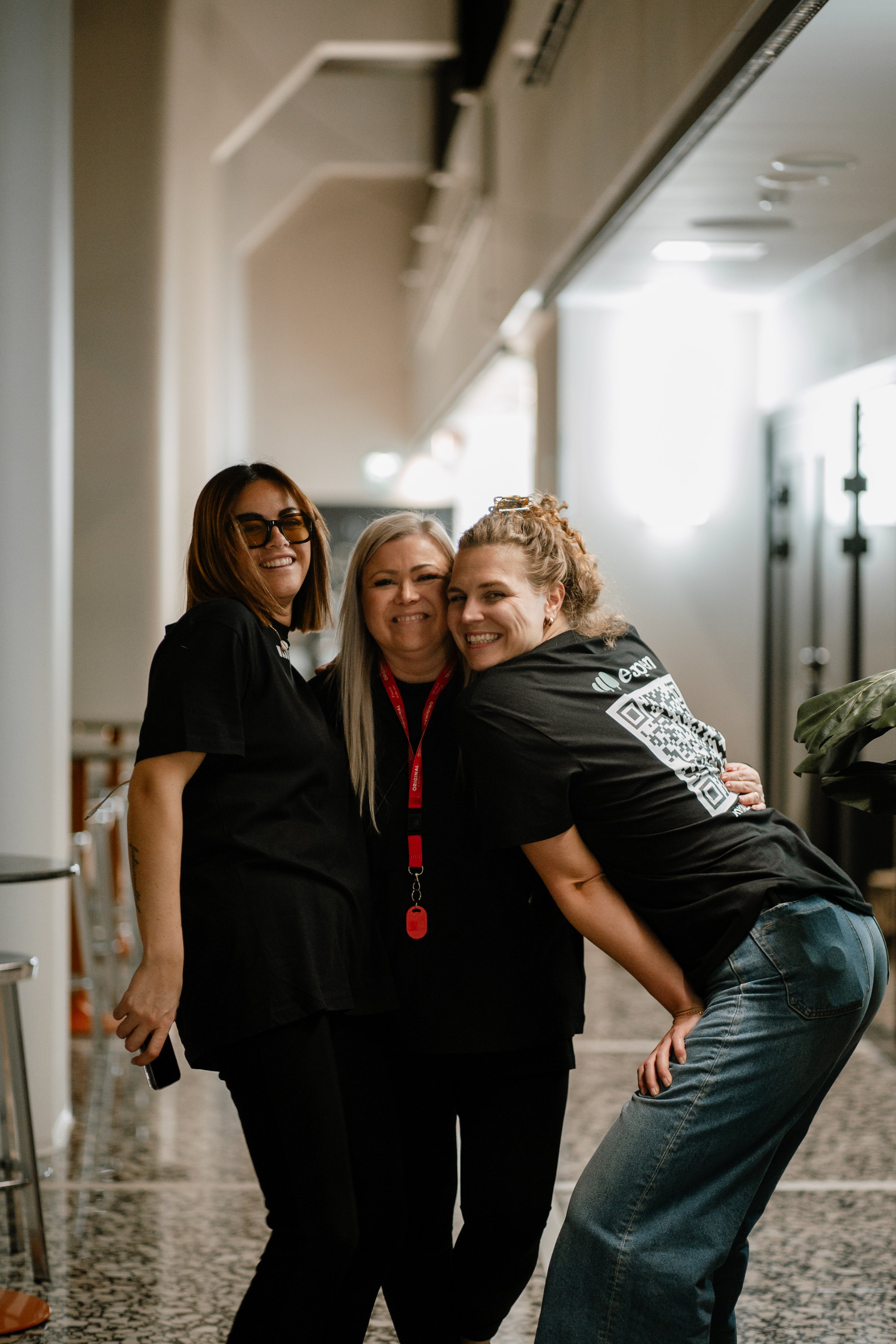 Three women standing close together and smiling, two of them hugging, in an indoor hallway with natural light.