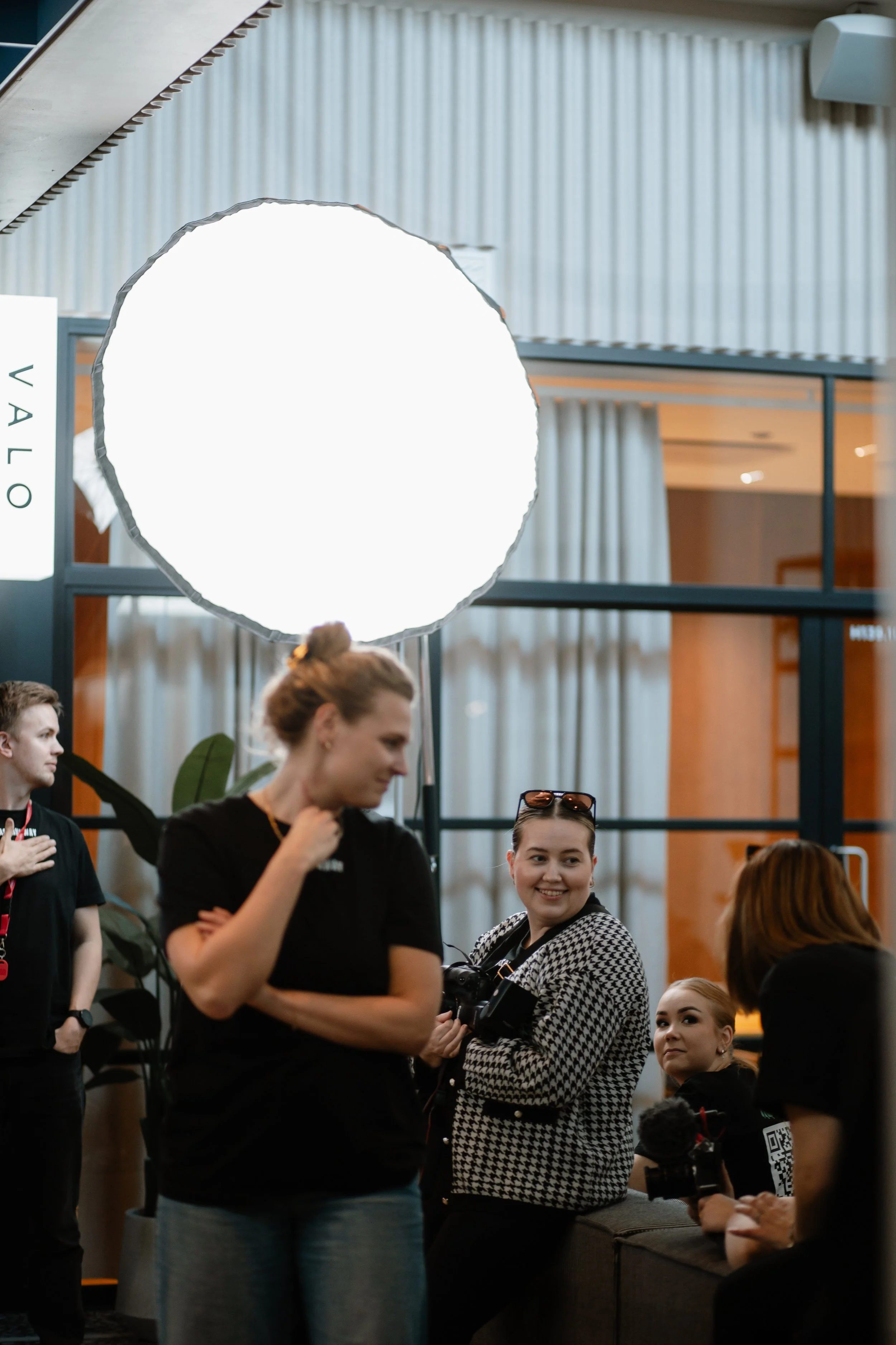 Women gathered in a modern indoor space with a large illuminated reflector overhead, engaged in conversation. One woman with glasses and a camera looks smiling at another woman, while others either chat or stand nearby. The background features glass partitions and plant decor.