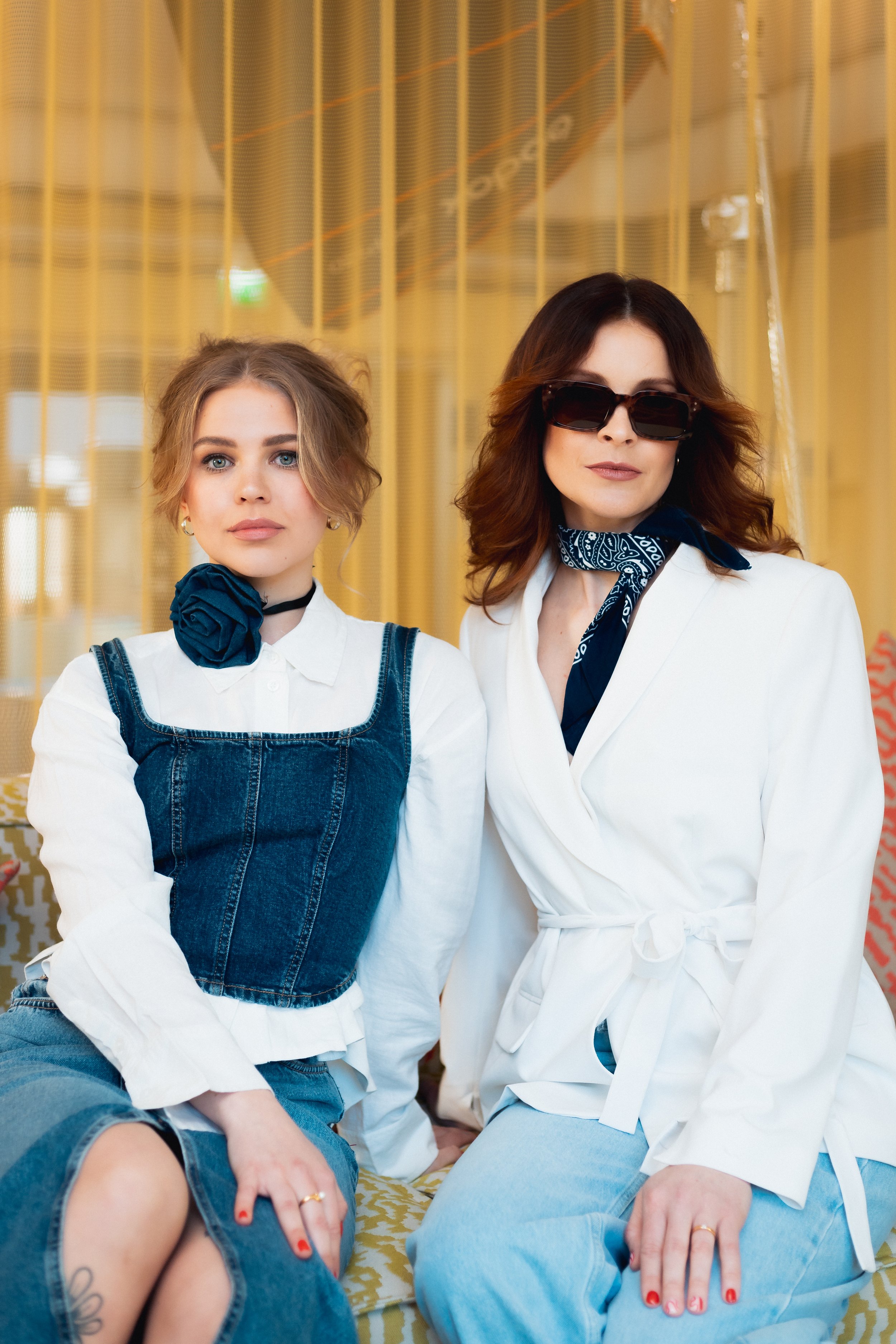 Two women sitting on a yellow patterned couch, one with short blonde hair wearing a white blouse with denim overalls, and the other with wavy brown hair wearing sunglasses, a white blazer, and a blue bandana around her neck. They are indoors with yellow curtains in the background.