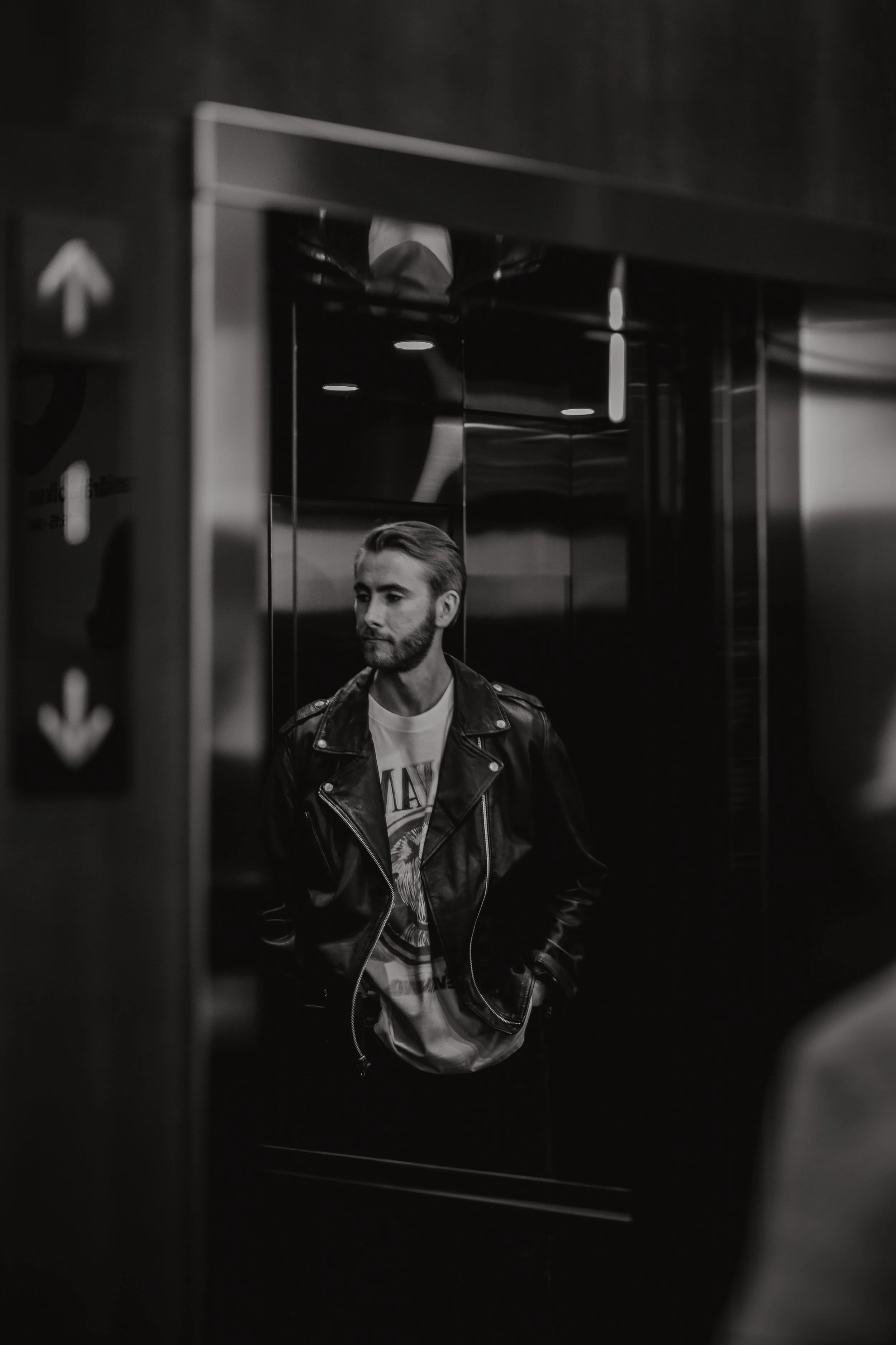 Black-and-white photo of a young man with beard, wearing a leather jacket and graphic t-shirt, standing inside an elevator, looking to the side.