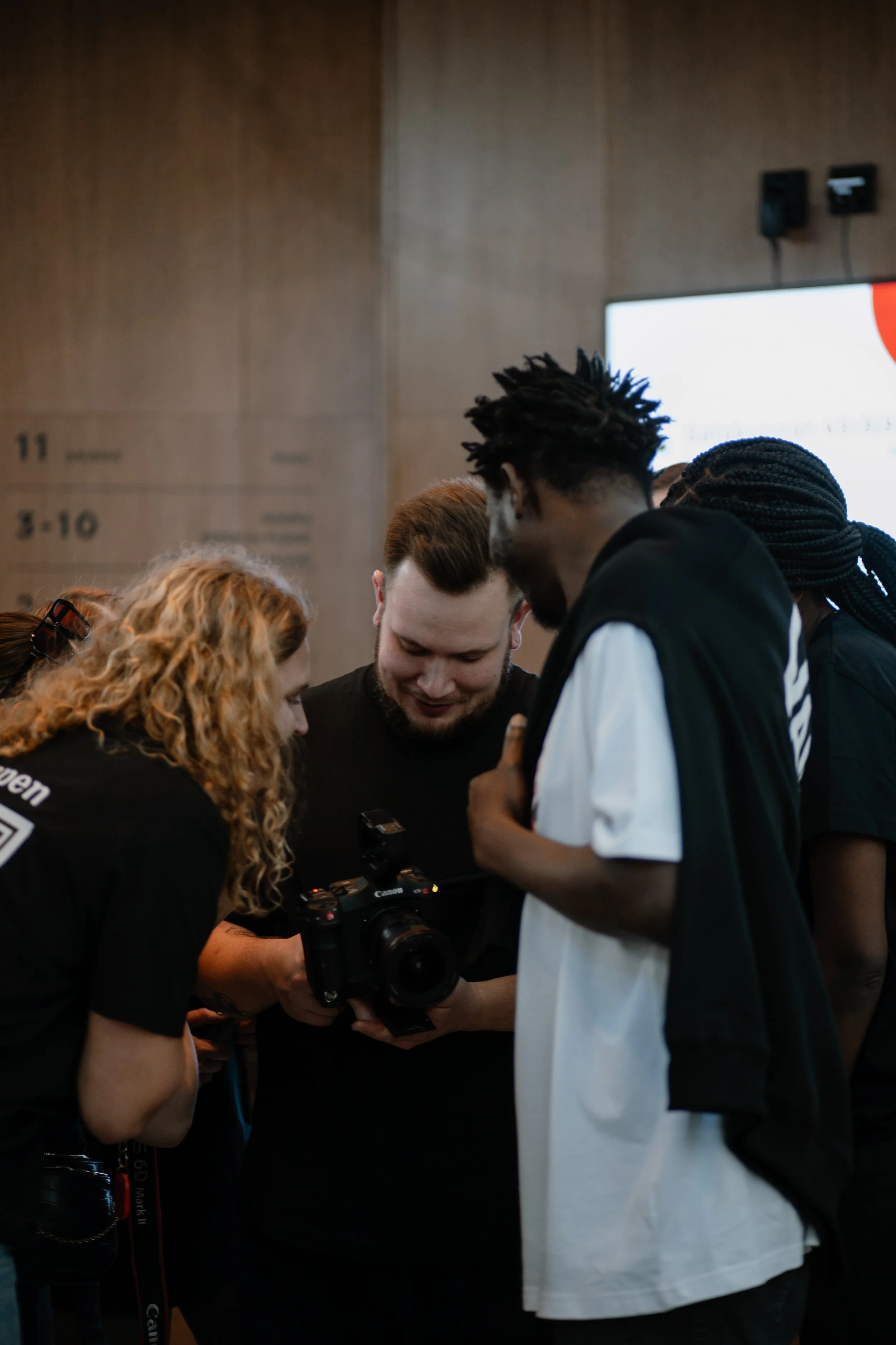 Group of four people gathered around a camera, looking at its screen, indoors with a wooden wall and a large screen in the background.