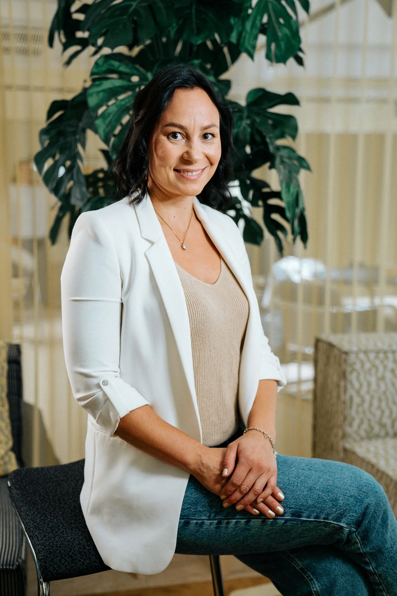 A woman with dark hair dressed in a white blazer, beige top, and jeans, sitting on a chair in front of a large green plant in an indoor setting, and smiling at the camera.