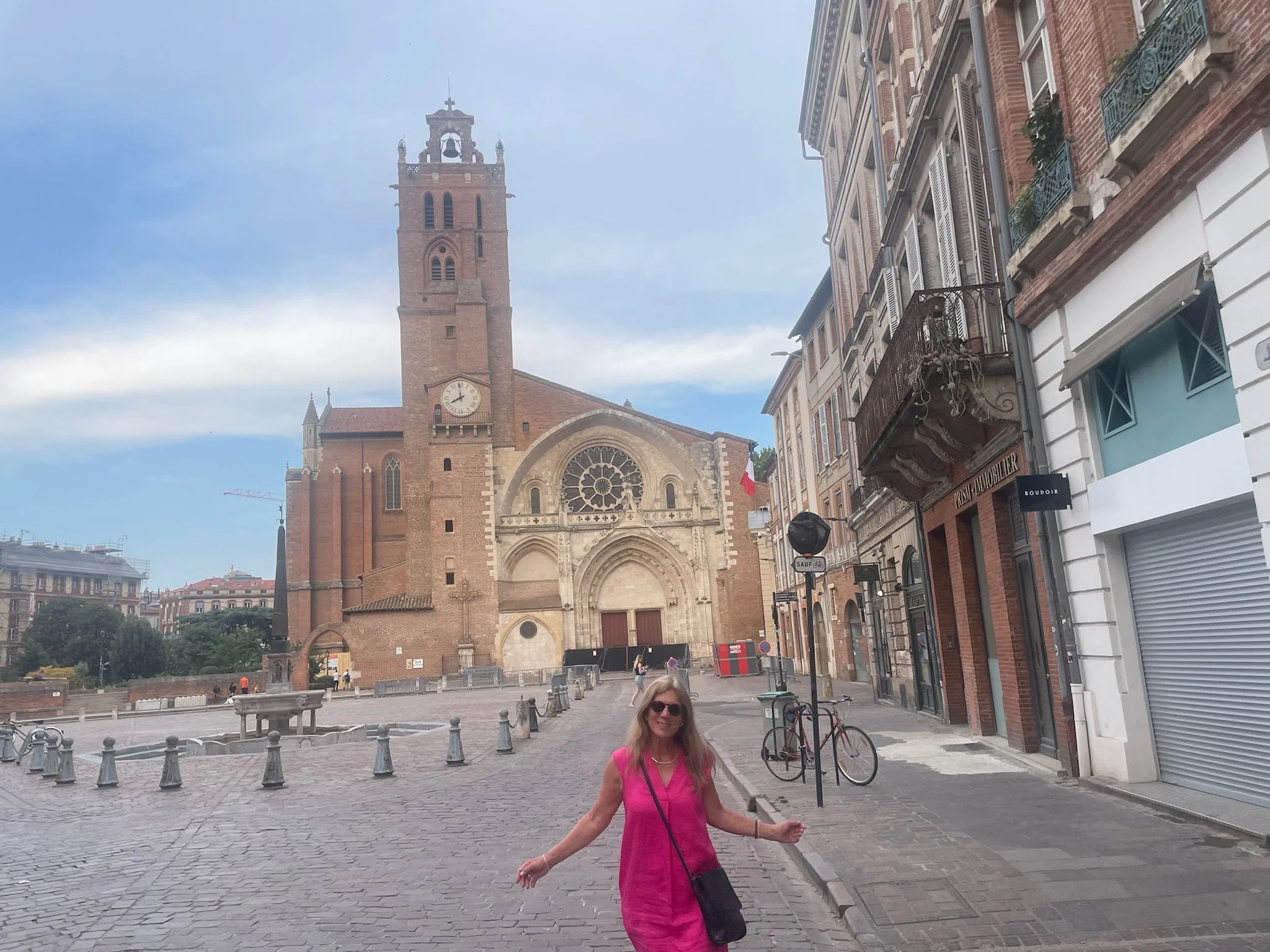 A woman in a pink dress walking on a cobblestone square in front of a historic church with a clock tower. There are bicycles, street signs, and buildings with balconies on the right side.