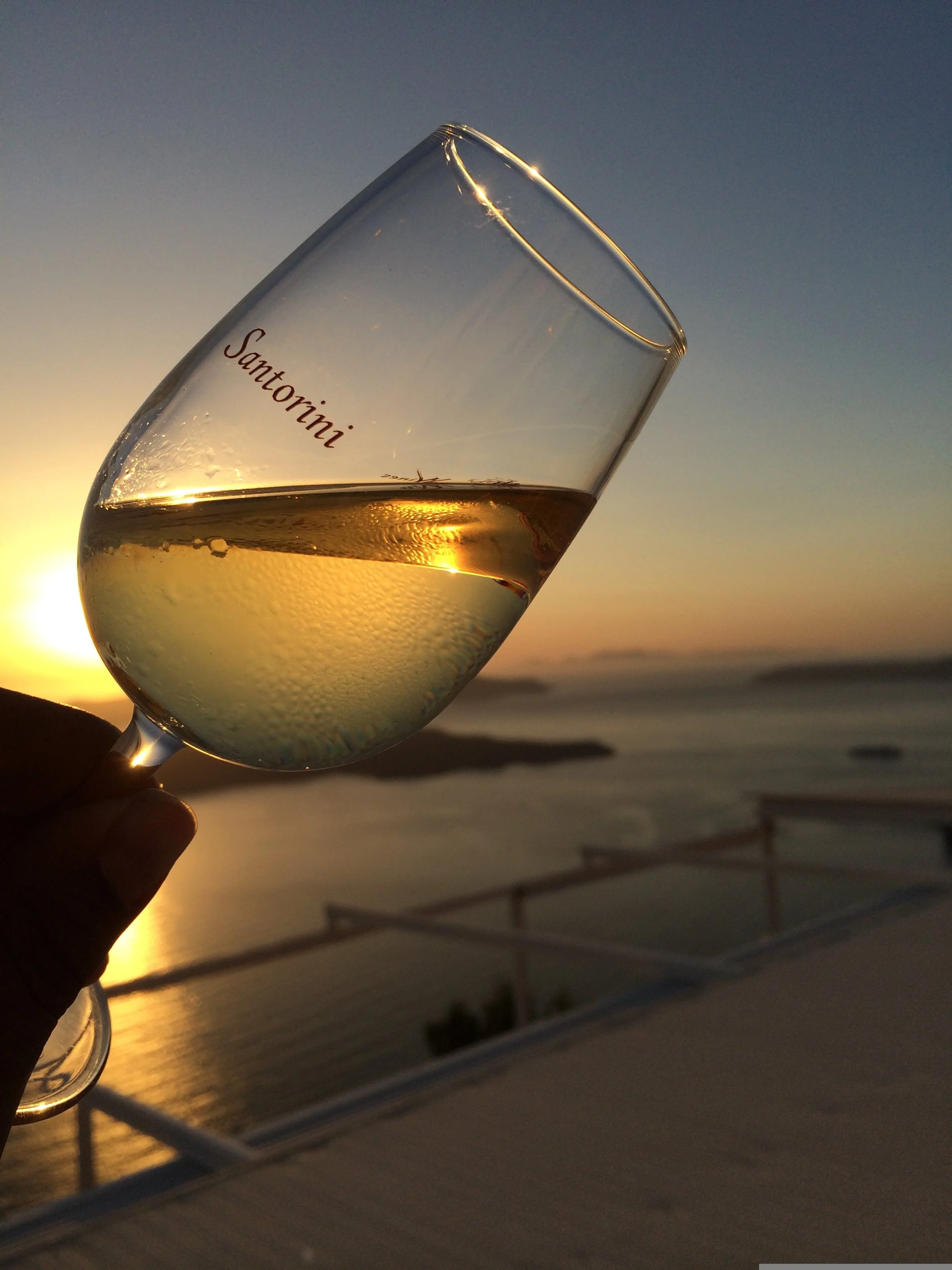 A glass of white wine held up against the backdrop of a sunset over the ocean, with beach umbrellas and lounge chairs in the distance.