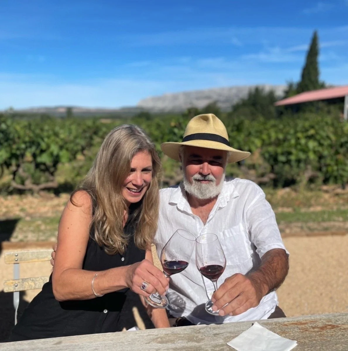 A woman and a man sitting outdoors, smiling, holding glasses of red wine, with vineyards and hills in the background on a sunny day.