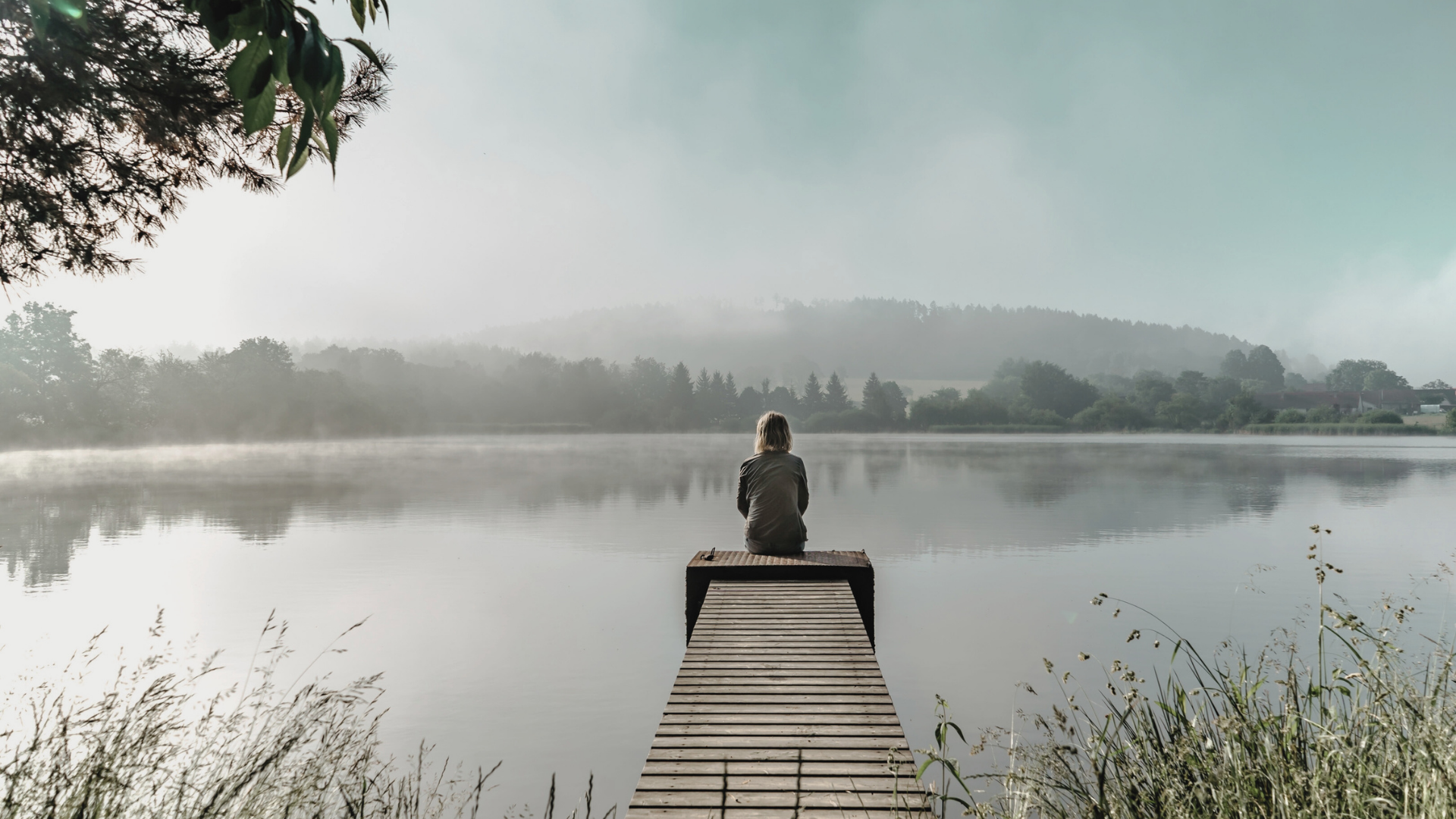 Person sitting on a dock by a lake, surrounded by fog and trees, with hills in the background.