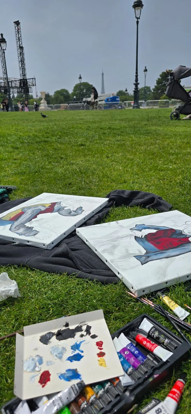 Paint supplies and two painted canvases depicting cartoon characters on the grass in a park, with Paris landmarks and people in the background.