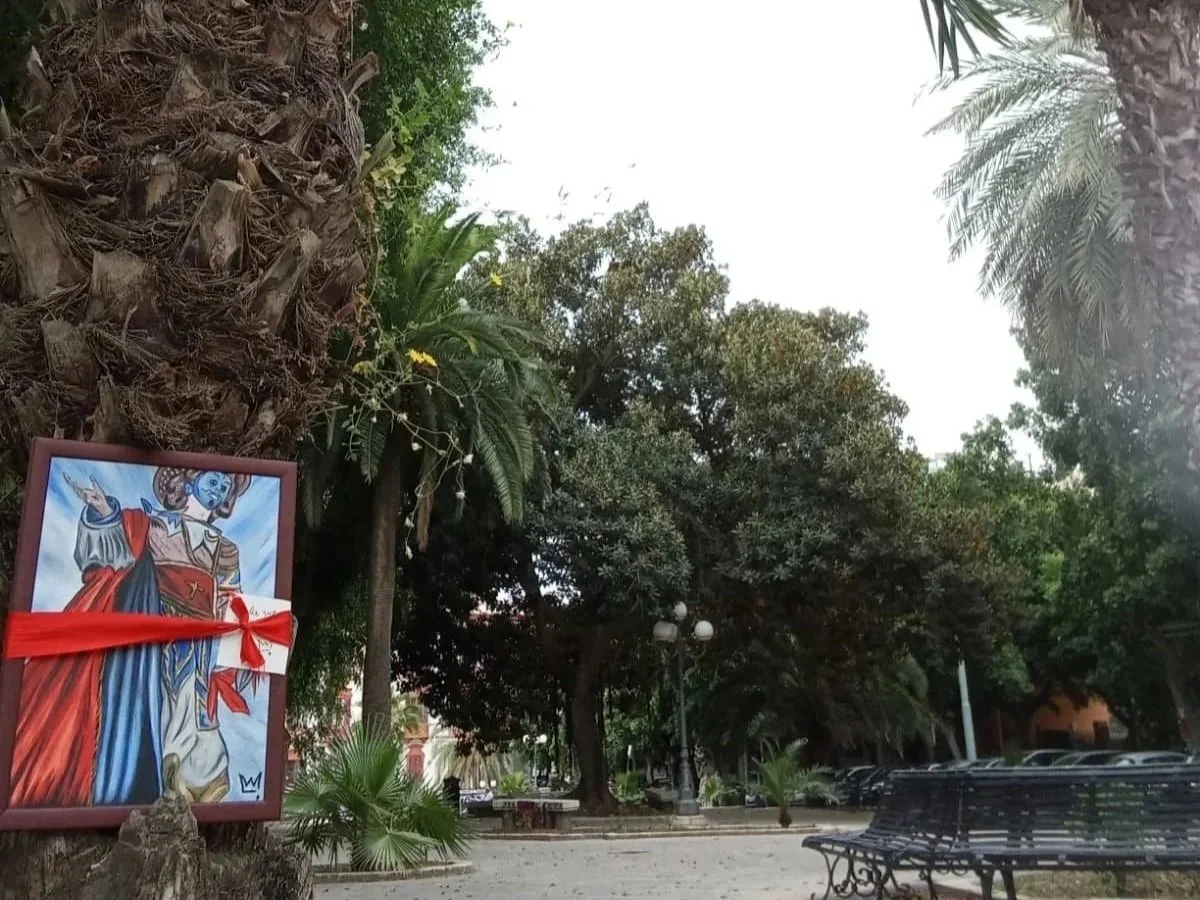 A religious image of Jesus Christ as a king with a red and blue cloak, a crown, and a red ribbon across the front, mounted on a tree trunk in a park with benches, trees, and a parking lot in the background.
