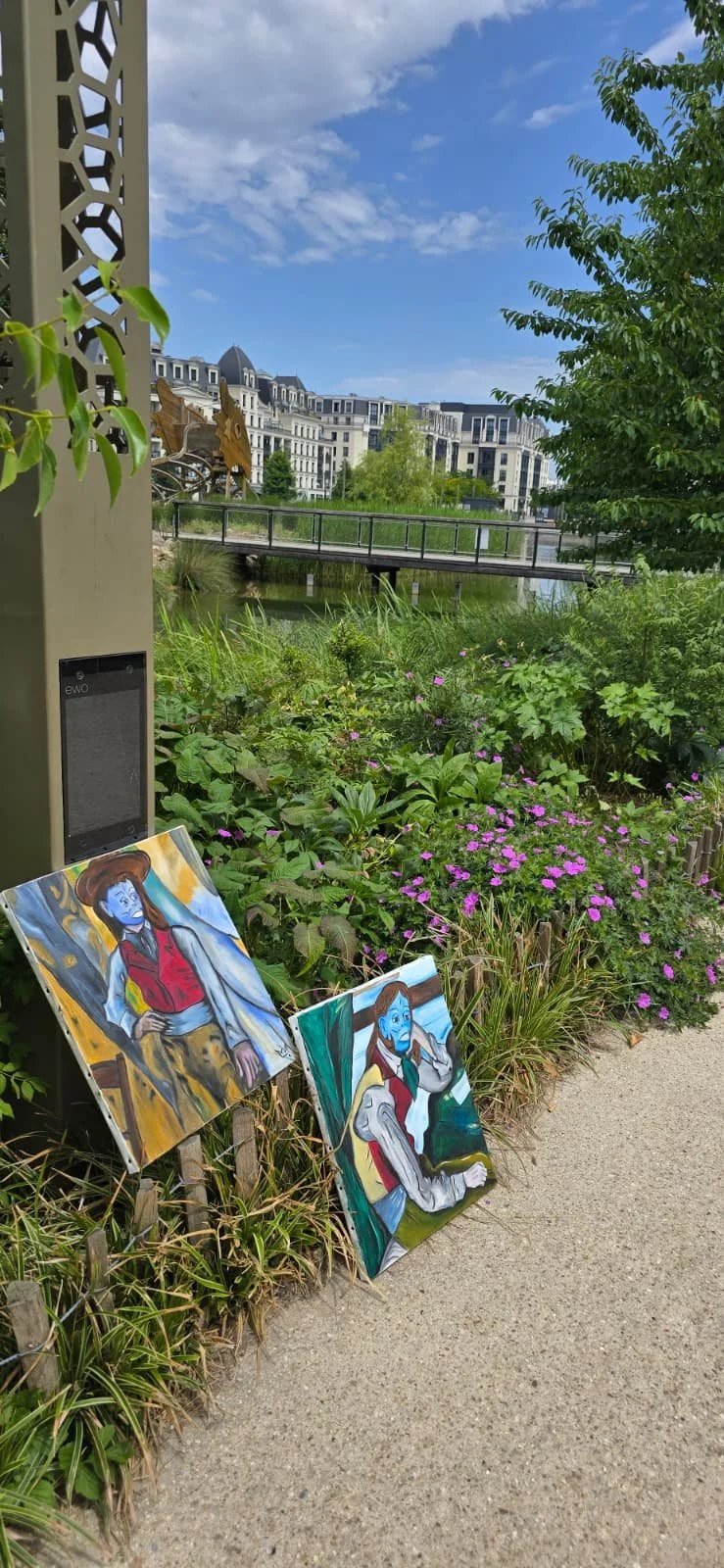 Two colorful paintings of a blue-skinned woman in red clothing resembling a character are displayed on the ground near green plants and pink flowers, with a backdrop of modern buildings, a bridge, and a blue sky with clouds.