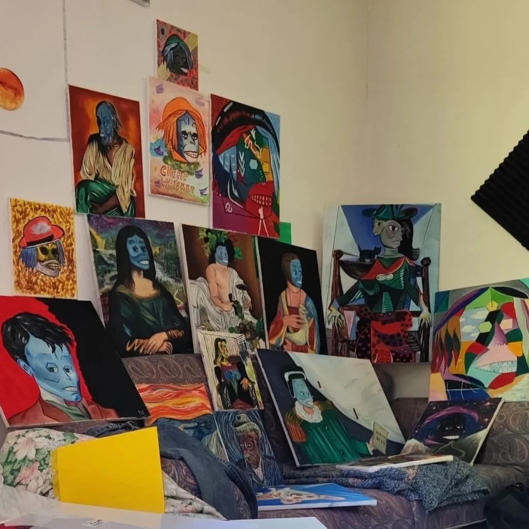 Wall and sofa covered with colorful paintings, including portraits and abstract art, with some leaning on the sofa and others hanging on the wall. The paintings depict various characters and styles, some with blue faces and vibrant backgrounds.