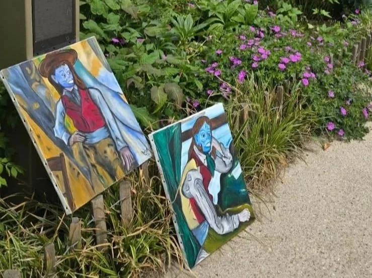 Two colorful paintings on easels next to a garden with pink flowers and green foliage. The paintings depict people with blue faces.