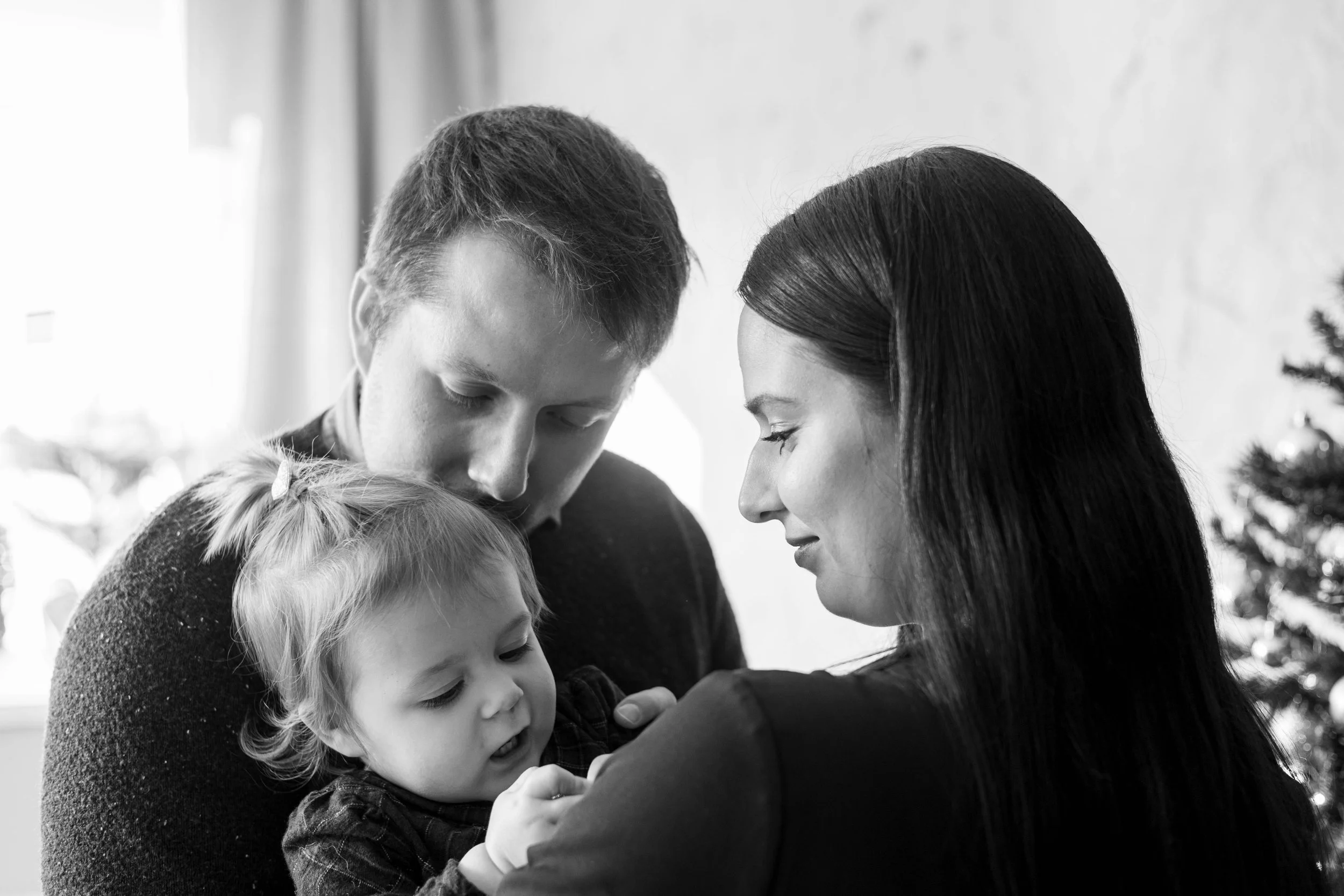 A black and white photo of a man and woman looking lovingly at a young girl, who appears to be their daughter, as she looks at something in her hand, with a Christmas tree in the background.