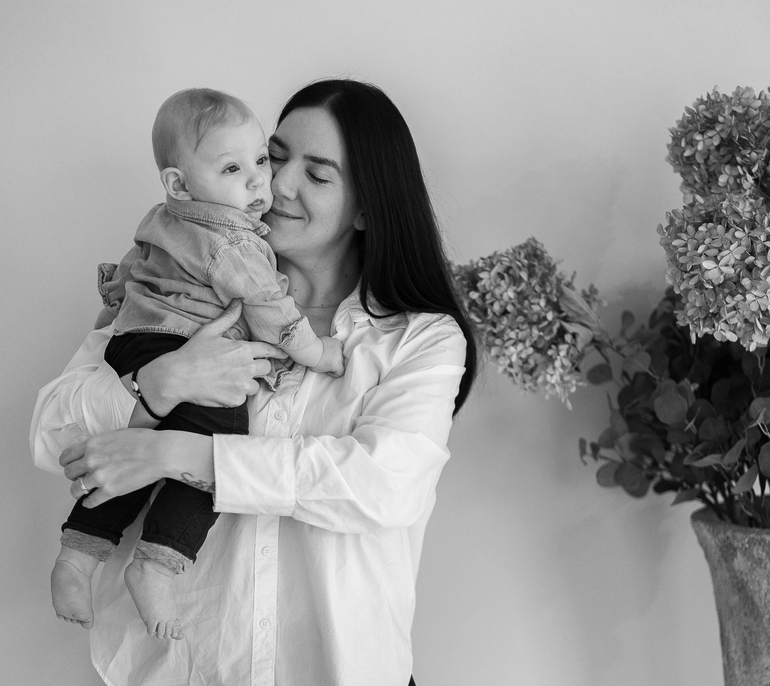 A woman with long dark hair holding a young child, both with closed eyes and smiling, in front of a plain wall with a vase of flowers to the side.