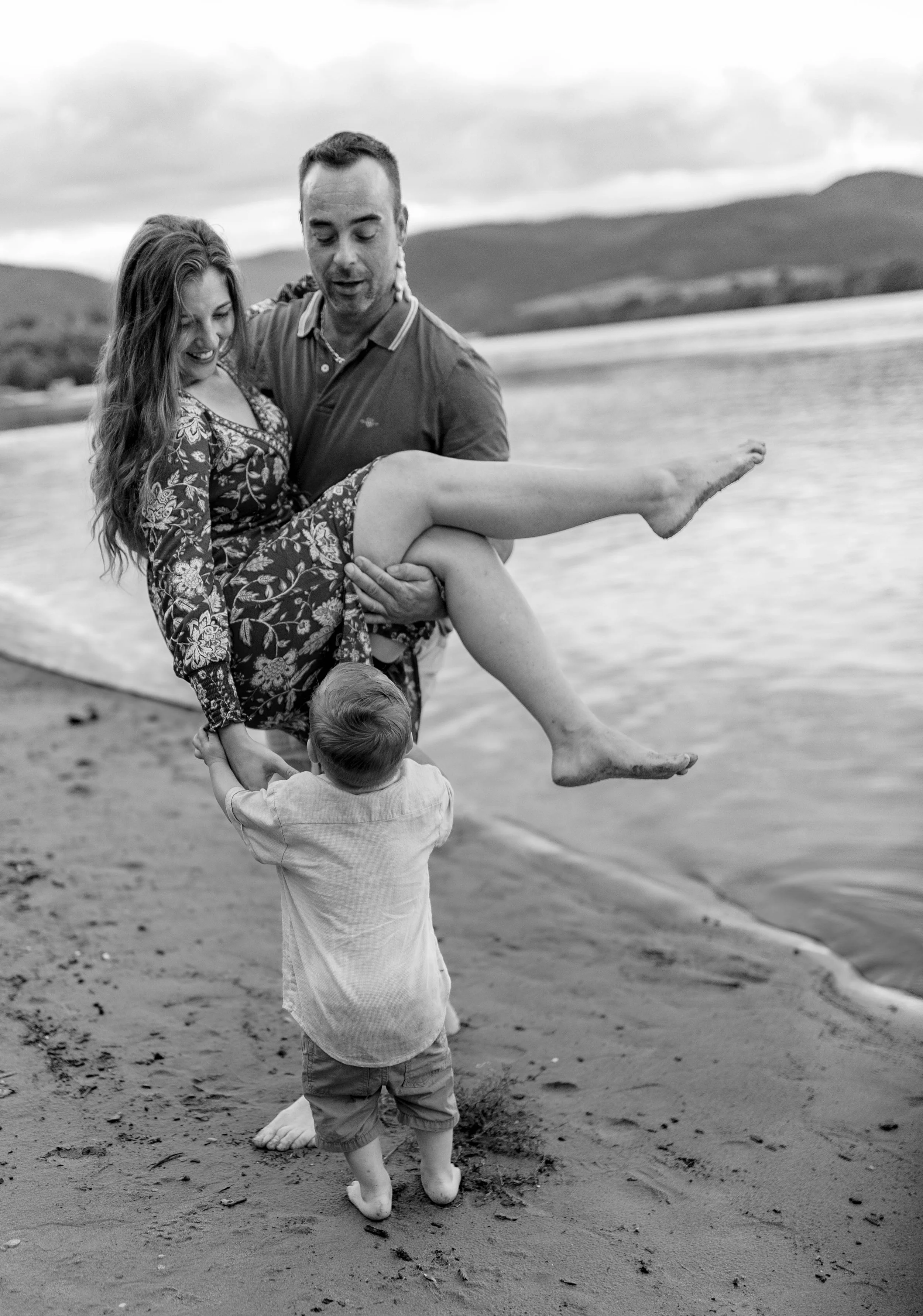 A man carrying a woman on his arms on a beach with a young boy holding her legs. The woman is smiling, and the boy is looking up at them. The scene is outdoors near a body of water with hills in the background.