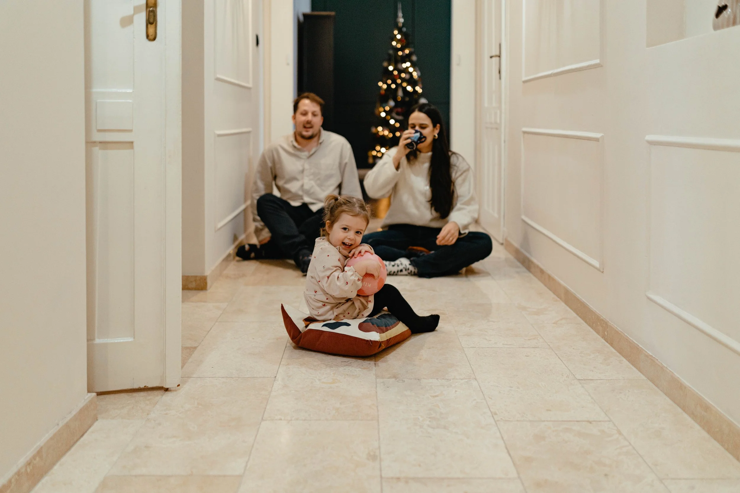 A black and white photo of a man and woman looking lovingly at a young girl, who appears to be their daughter, as she looks at something in her hand, with a Christmas tree in the background.