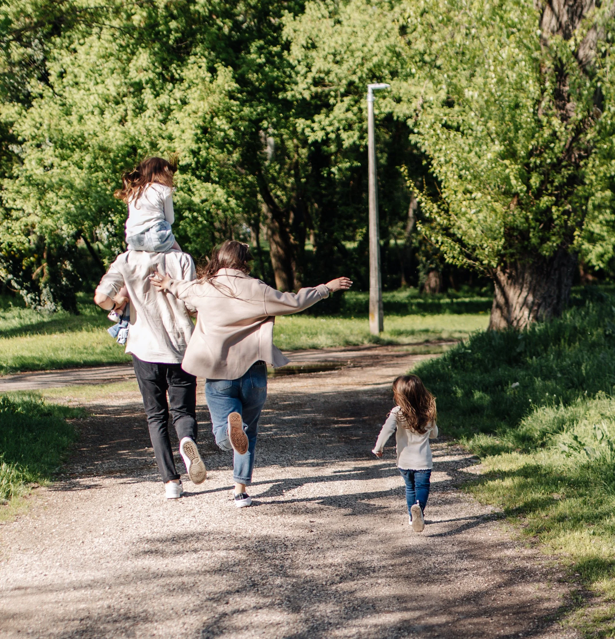 A family of four walking on a dirt path in a park with lush green trees, with a streetlamp in the background, on a sunny day.