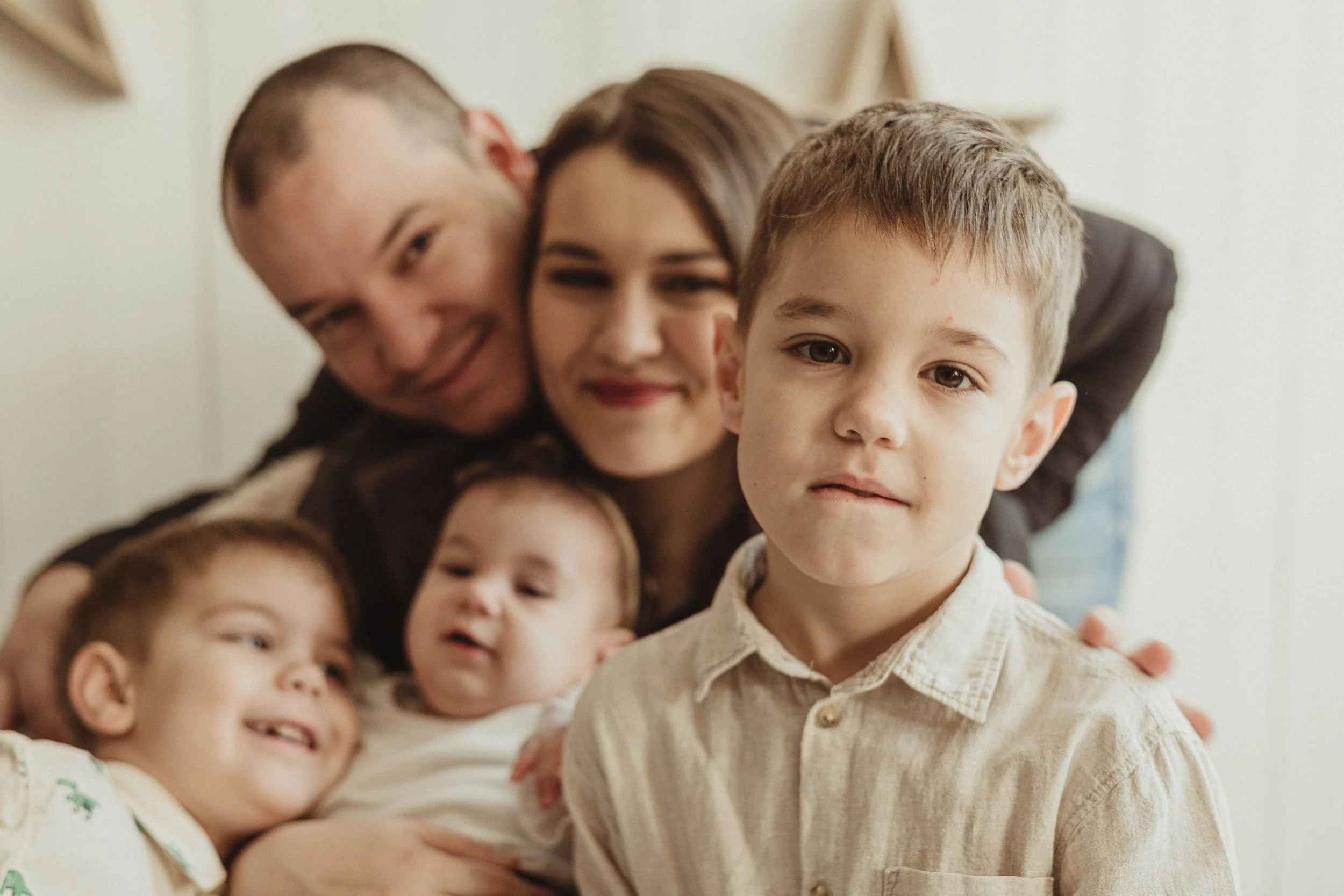 A family of five, including two young children, a toddler, a young boy, a woman, and a man, posing closely together indoors, with the boy in the front and the others smiling behind him.