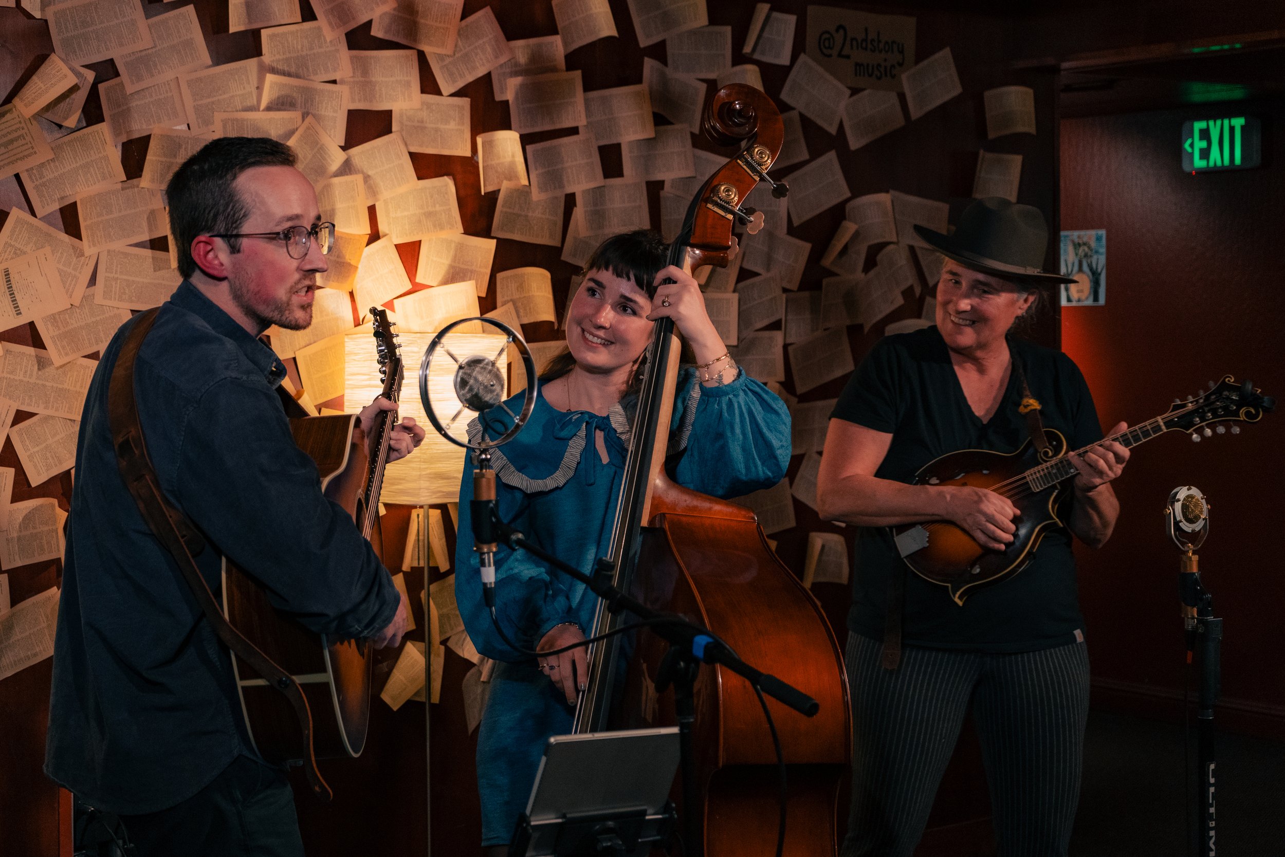 Three musicians performing in a cozy indoor setting with walls decorated with open books. The man on the left plays an acoustic guitar, the woman in the middle plays a double bass, and the woman on the right plays a mandolin. They are smiling and app