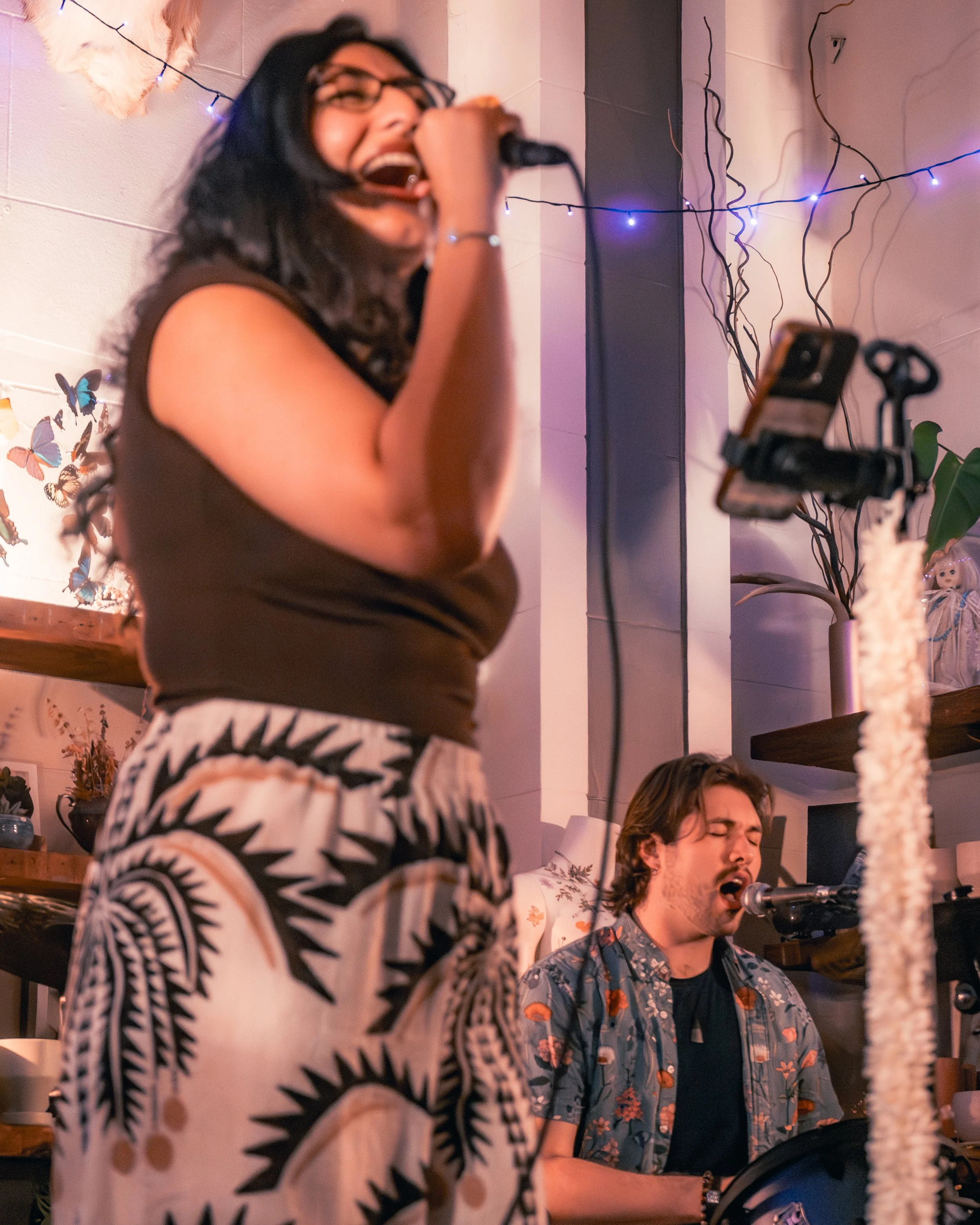 A woman singing into a microphone with a man playing a drum, both appear to be performing or jamming together in a cozy, decorated room with fairy lights and butterflies on the wall.