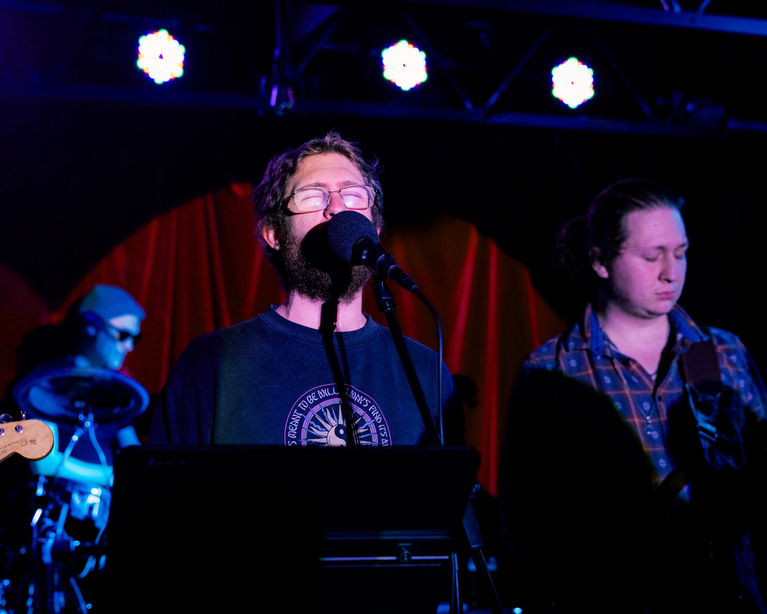 Three musicians performing on stage with red curtain background and stage lights, one singing into a microphone with glasses, a female musician with closed eyes, and a drummer wearing glasses and a Santa hat.